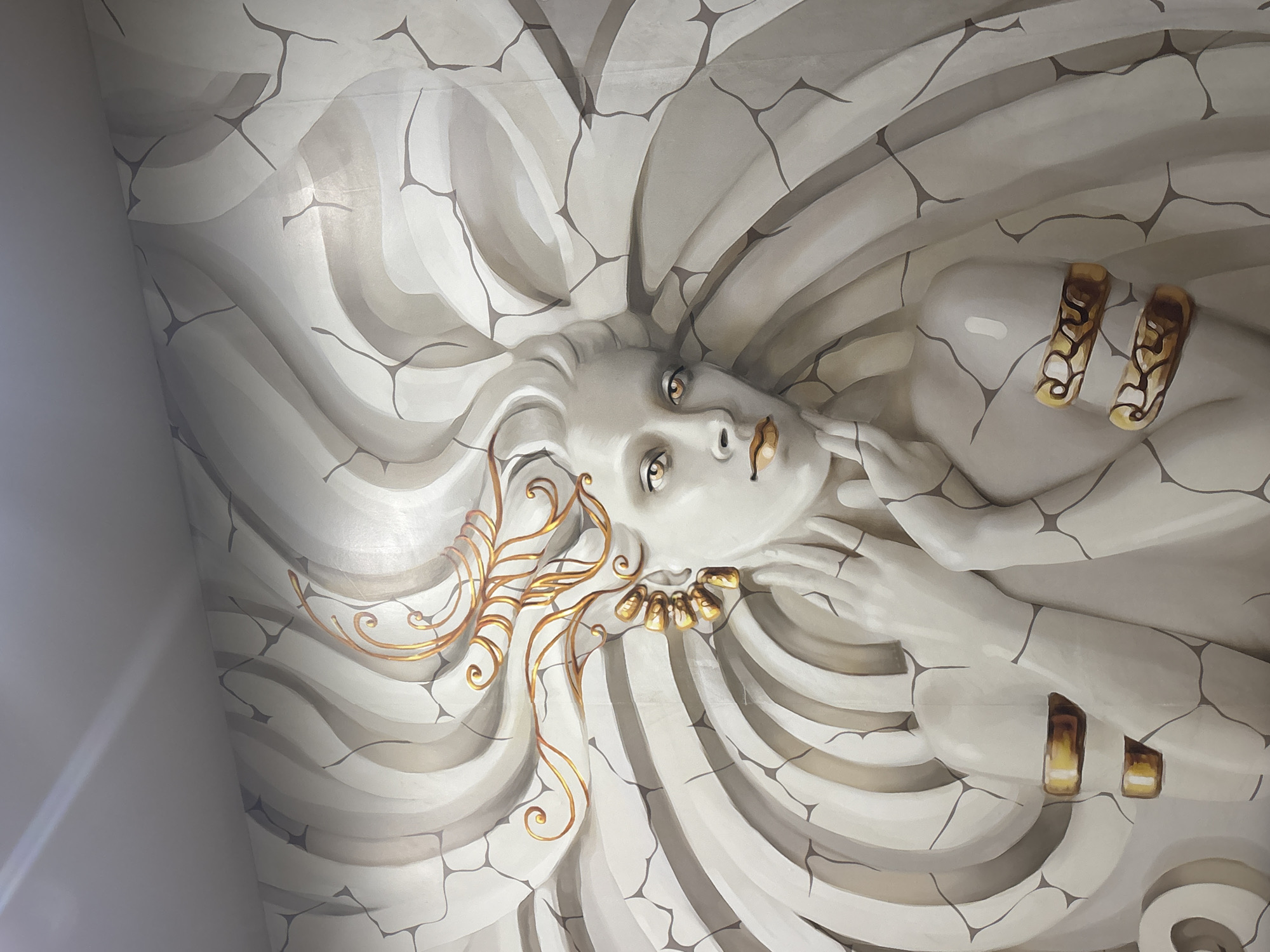 The image displays a large-scale, stylized mural or relief artwork, likely on a ceiling or wall. The central subject is a female figure depicted from the shoulders up, with her head slightly tilted and her gaze directed upwards and to the viewer's right. Her eyes are a striking amber-brown, and her lips are painted gold.
Her hair is rendered as voluminous, flowing waves of grey and white, seamlessly blending with the background. Intricate golden, swirling patterns accentuate the hair on the left side of her face, and she wears a golden necklace around her throat. Her right arm is visible, bent at the elbow, with her hand gently resting near her face. Two golden, patterned cuff-like armbands adorn her right forearm.
The artwork's palette is predominantly monochromatic, utilizing shades of white, light grey, and darker grey, with vibrant golden accents providing a focal point. A notable detail is the pervasive crack-like pattern of dark lines that covers both the figure and the background, giving the impression of aged stone or a shattered surface. The background consists of sweeping, wave-like formations in varying shades of grey, enhancing the sense of movement and depth.
The overall style evokes a blend of classical sculpture and mythological imagery, with the figure appearing almost ethereal yet rooted in a stone-like texture. No visible text is present in the image. This artwork is located in Troyan, Bulgaria.