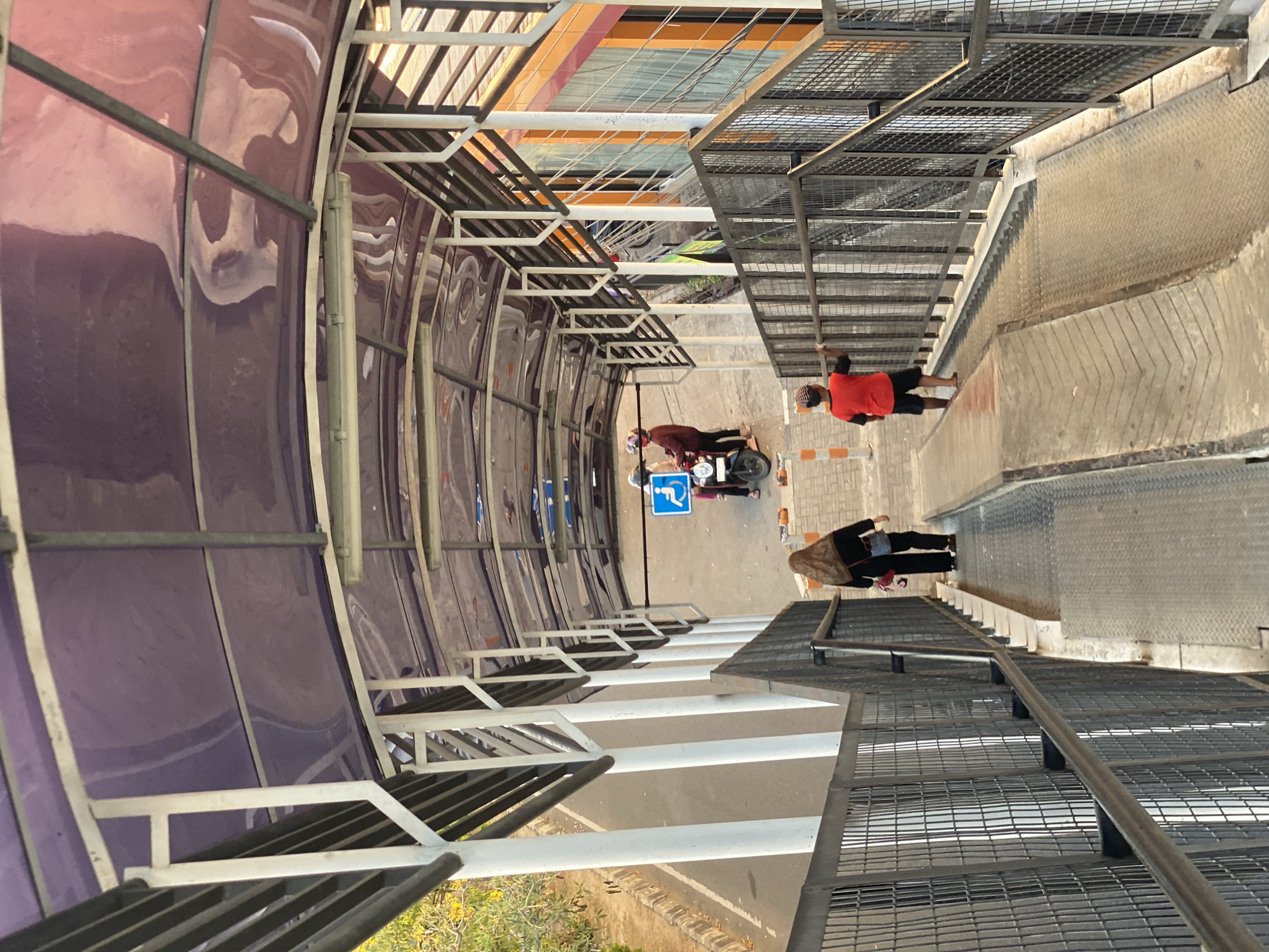 The image depicts an elevated pedestrian walkway with a purple translucent roof supported by white pillars and dark metal beams. The walkway has metal grating on the sides and is equipped with ramps and stairs. A person wearing a headscarf and black clothing is walking down the ramp, carrying a red bag. Another person, a child wearing a red shirt and black shorts, is also walking down the ramp, holding onto the railing. Further ahead, two individuals on a motorcycle are visible near a blue sign indicating accessibility for persons with disabilities. The walkway is adjacent to a road with a sidewalk and some vegetation. The building to the right of the walkway is painted in orange and yellow. The overall scene suggests a public transportation hub or a pedestrian crossing in Banjarbaru, Indonesia, likely near a road or train station, given the presence of the motorcycle and the accessible pathway.