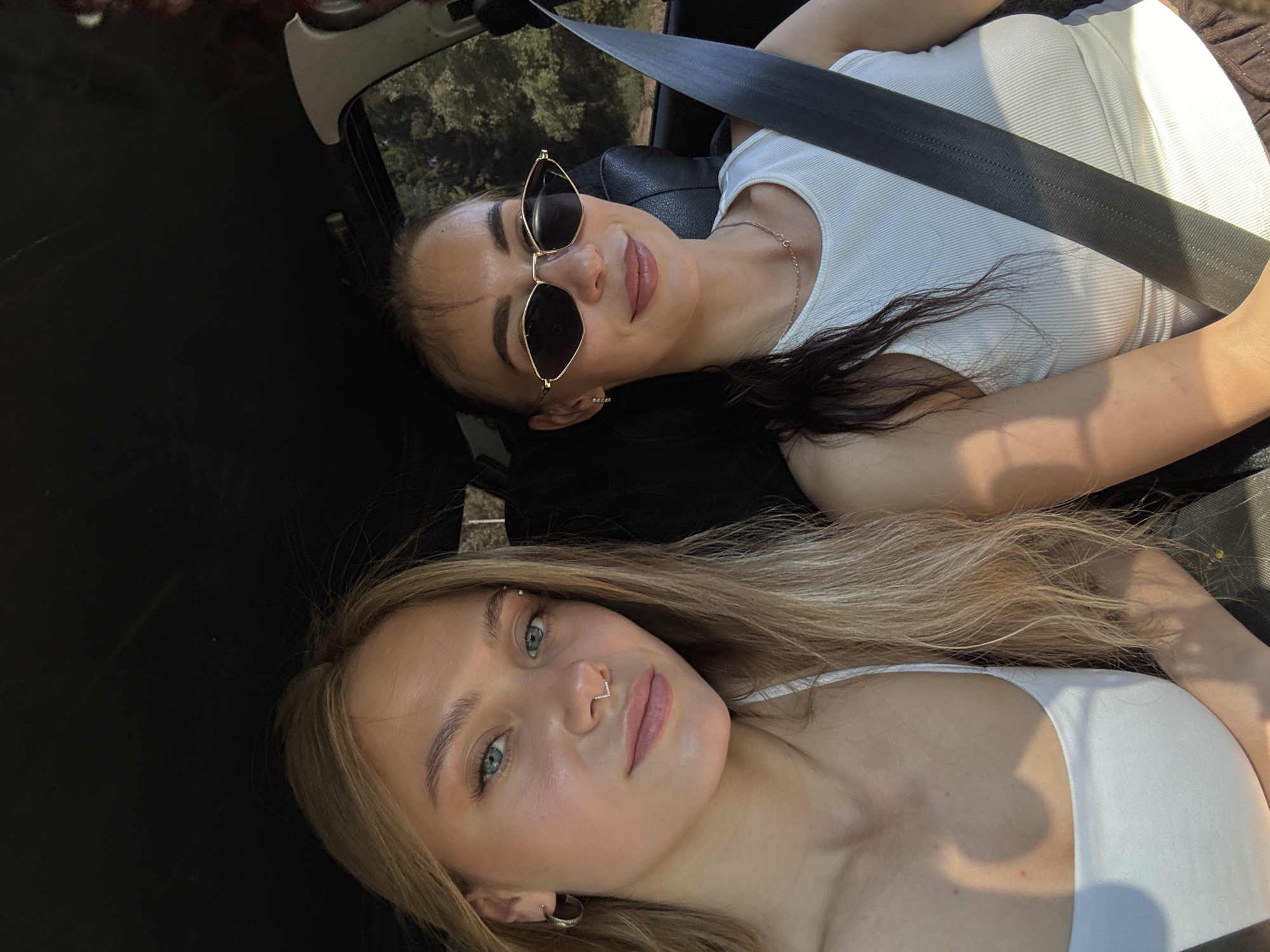 Two young women are inside a vehicle, appearing to be on a road trip, with the camera capturing a selfie. The woman on the left has light blonde hair, blue eyes, and is wearing a white tank top. She has a nose ring with a septum piercing and a hoop earring in her left ear. Her hair is slightly windswept. The woman on the right has dark brown hair, wears stylish, angular sunglasses, and a white ribbed tank top. She has a delicate necklace around her neck and small earrings. Both women are smiling. The interior of the car is visible, with the headliner dark and a seatbelt prominently displayed across the woman on the right. Sunlight streams in from the windows, casting shadows and highlighting their faces. Through the windows, a blur of green foliage and trees can be seen, suggesting a rural or natural setting outside the car. The specific location in Chaikovsky, Russia, cannot be determined from the visual information provided.