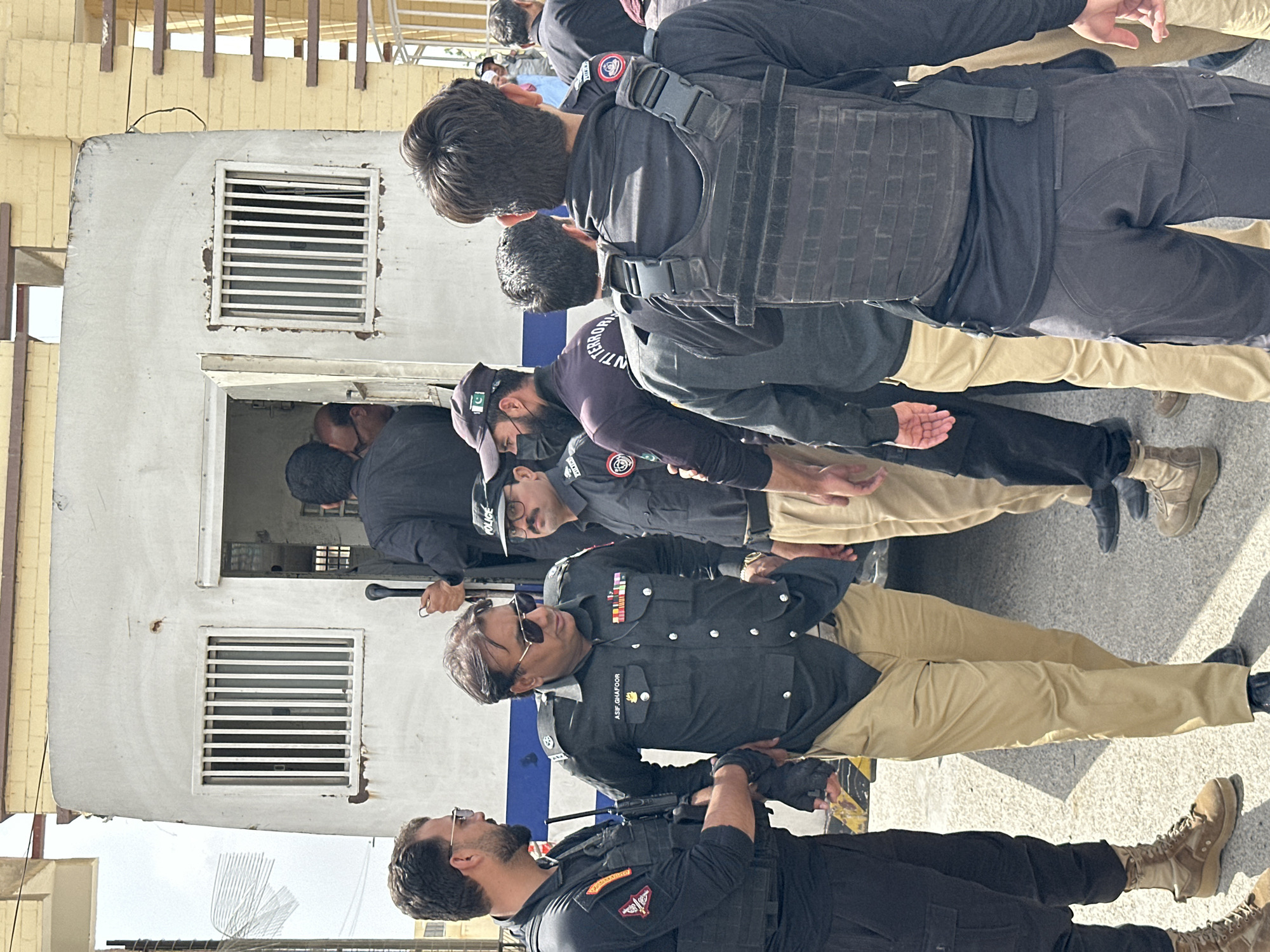 In an outdoor scene in an unknown city, a group of police officers surround a white van with barred windows, possibly a prisoner transport vehicle. Some officers are in uniform, while others wear tactical gear. One officer, identified by the name "ASIF GHAFFOOR" on his uniform and wearing sunglasses, appears to be in charge, talking to another uniformed officer. The environment suggests a bright, sunny day with a building made of yellow brick in the background. The overall impression is one of security and law enforcement activity.