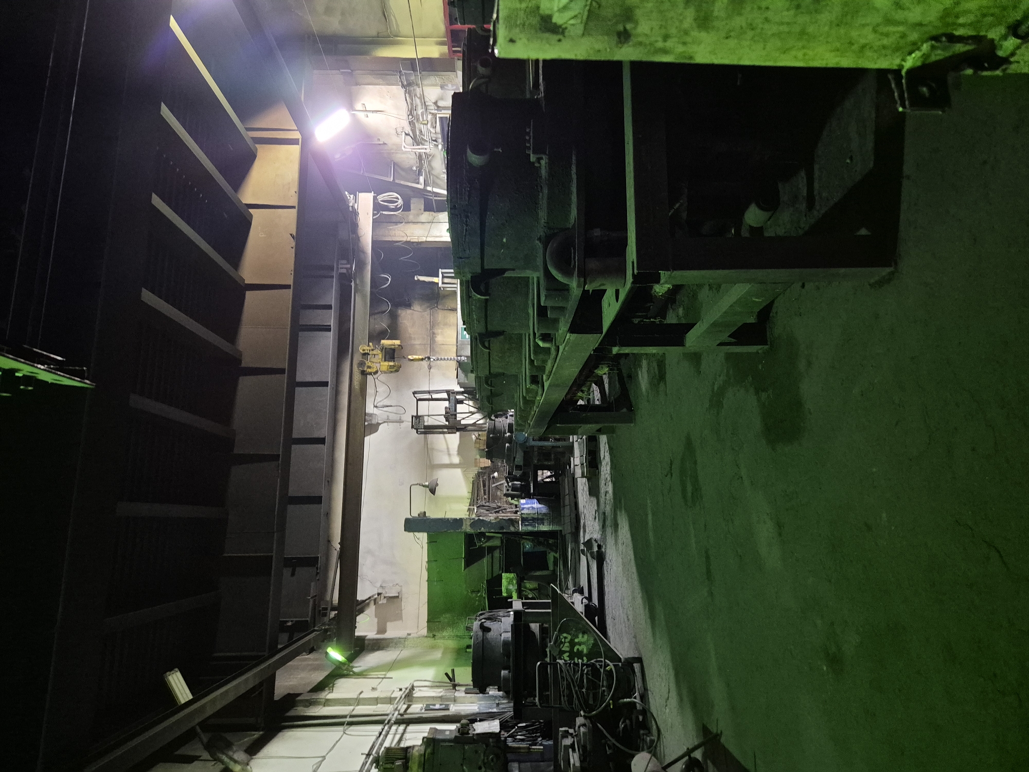 The image depicts the interior of a dimly lit industrial workshop in Pavlodar, Kazakhstan. The scene is dominated by large, dark machinery and equipment, casting long shadows across the concrete floor. The lighting appears to be artificial, with some areas illuminated by harsh, cool-toned lights, while others are shrouded in darkness. The walls are bare and appear to be concrete or stucco, with some sections painted a faded green. The ceiling is high, with exposed beams and ductwork, suggesting a functional rather than aesthetic design. Various industrial components, such as molds or large metal casings, are arranged on workbenches and the floor. The overall impression is one of a working industrial space, possibly a foundry or manufacturing facility, with a utilitarian and somewhat neglected appearance. There are no people visible in the image.