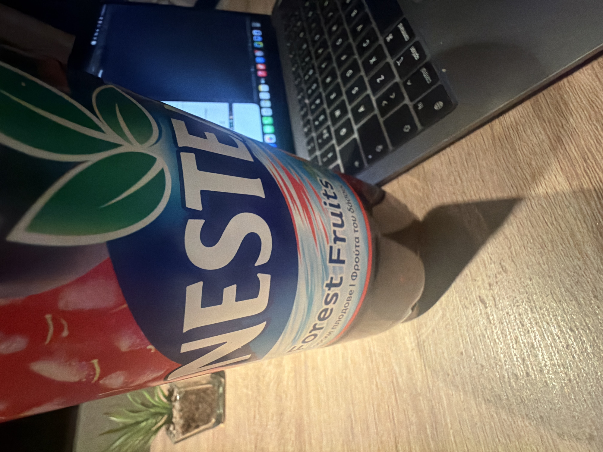 An image captured from a slightly elevated, rotated perspective displays a casual indoor setting, likely in Plovdiv, Bulgaria, given the linguistic cues. The primary subject is a plastic bottle of NESTE iced tea, lying horizontally on a wooden surface. The label prominently features the brand name "NESTE" and the flavor "Forest Fruits". Below the English text, the flavor is also written in Bulgarian ("Диви плодове") and Greek ("Φρούτα του δάσους").
Behind the bottle, a silver laptop, likely a MacBook, is open on the same wooden surface. Its keyboard, with visible keys such as "option", "command", "X", "C", and "V", is partially visible, indicating an active workspace. The laptop screen is illuminated, showing various application icons and what appears to be an open interface, suggesting it's in use.
The overall lighting in the scene is dim, with the laptop screen providing the main source of light, suggesting it is evening or nighttime. Shadows are cast on the wooden table from the bottle and potentially other unseen objects. A small potted plant with green leaves in a clear glass container is visible in the lower left background, adding a natural element to the otherwise tech-focused setup. The wooden surface has a distinct grain, and the shadows indicate a light source from above and to the left. No people are visible.