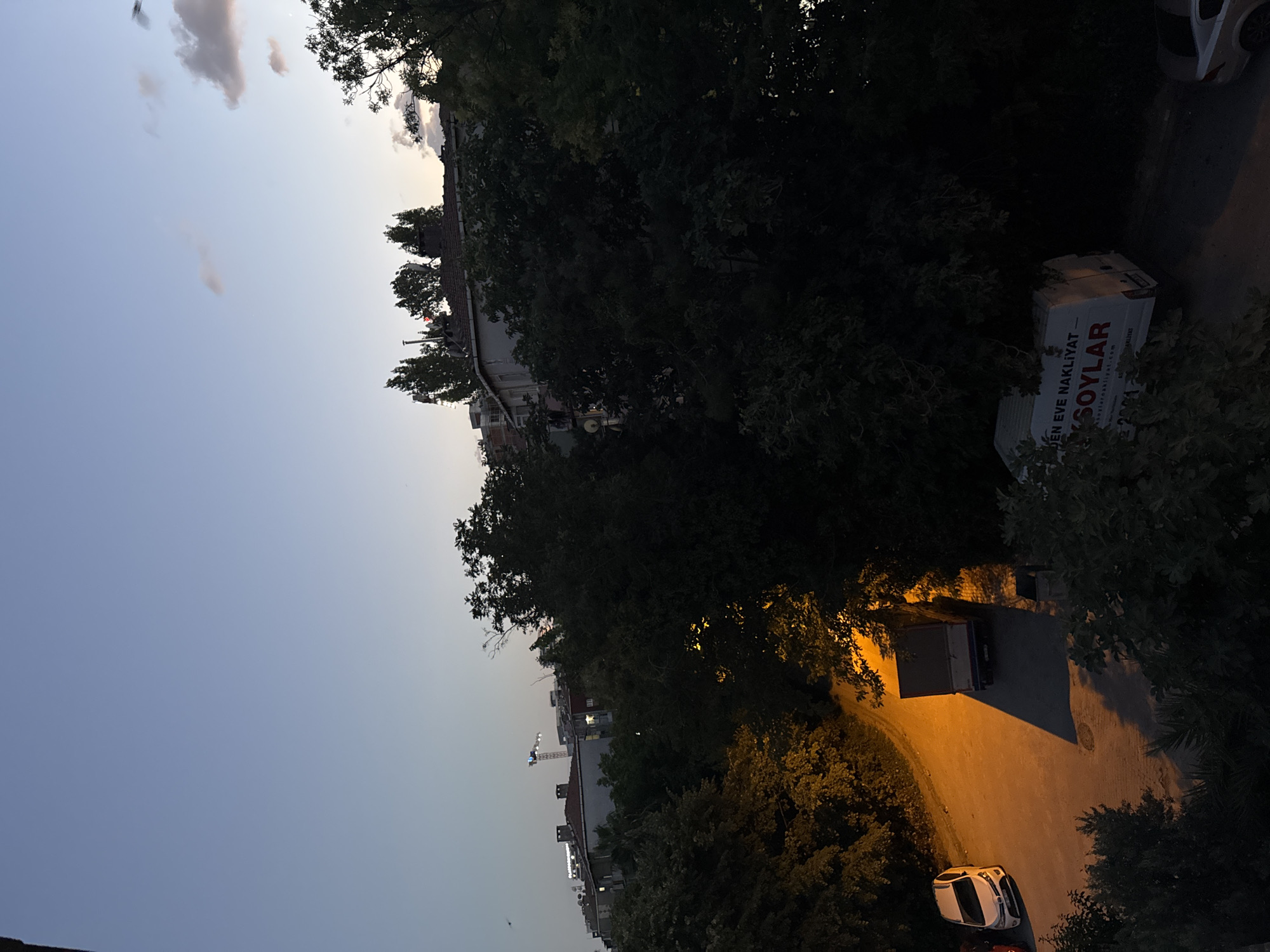 The image captures a twilight scene in Gungoren, Turkey, looking across a street lined with trees and apartment buildings. In the foreground, dense, dark green foliage dominates the lower portion of the frame, with a white car parked on the left side of a dimly lit, orange-hued street. Further down the street, a light-colored box truck is parked. Behind the street and trees, apartment buildings with visible windows and rooftop structures are visible. A construction crane is present in the distance, silhouetted against the pale blue and dusky orange sky. A few wispy clouds are scattered across the sky. The overall impression is an urban residential area during the transition from day to night.