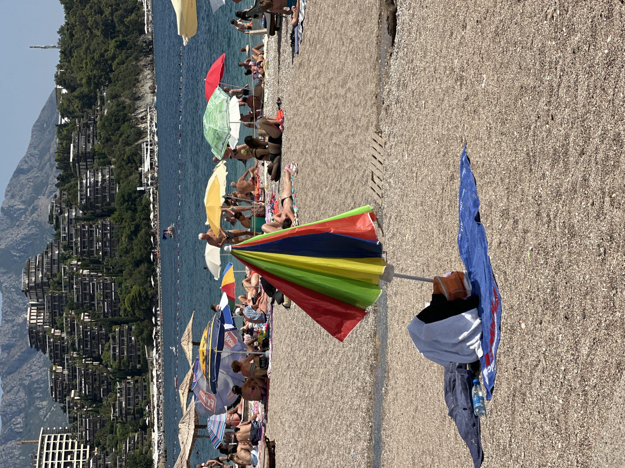 The image captures a sun-drenched beach scene in Budva, Montenegro, with the distinctive tiered architecture of the nearby hills forming the backdrop. In the foreground, a vibrant, multi-colored beach umbrella is prominently displayed, its segments of red, yellow, green, and blue standing out against the pebbled shore. Scattered on a blue towel behind the umbrella are various items of clothing, including a dark blue shirt and a white towel, alongside a partially visible orange bag and a plastic water bottle.
Further back, the beach is populated with numerous beachgoers, mostly seated or lying on towels under a variety of beach umbrellas. These umbrellas, in shades of beige, yellow, green, and red, provide shade for individuals and small groups. The sea, a deep blue, stretches out behind the beach, with a few people visible wading or swimming. The coastline is lined with more umbrellas and what appear to be lounge chairs, suggesting a well-established tourist destination.
The hilly terrain behind the beach is densely covered with lush green vegetation, interspersed with the characteristic terraced buildings that are a hallmark of Budva's landscape. These structures, appearing as multi-level hotels or apartment complexes, climb the hillsides, creating a striking contrast between the natural environment and the man-made structures. The overall impression is one of a busy, active beach on a clear, sunny day, with the picturesque Montenegrin coastline providing a scenic setting.