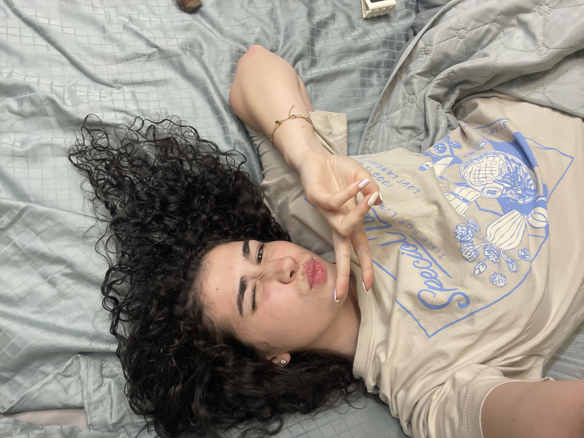 A young woman with curly hair is lying on a bed. She is wearing a light-colored t-shirt with a blue graphic. Her right arm is raised, with her fingers forming a peace sign. The bed is covered with a gray sheet and a gray quilted blanket is partially covering the sheet. The location of this image is likely not in Sofia, Bulgaria as there is no evidence of a city or Bulgarian influence within the image.