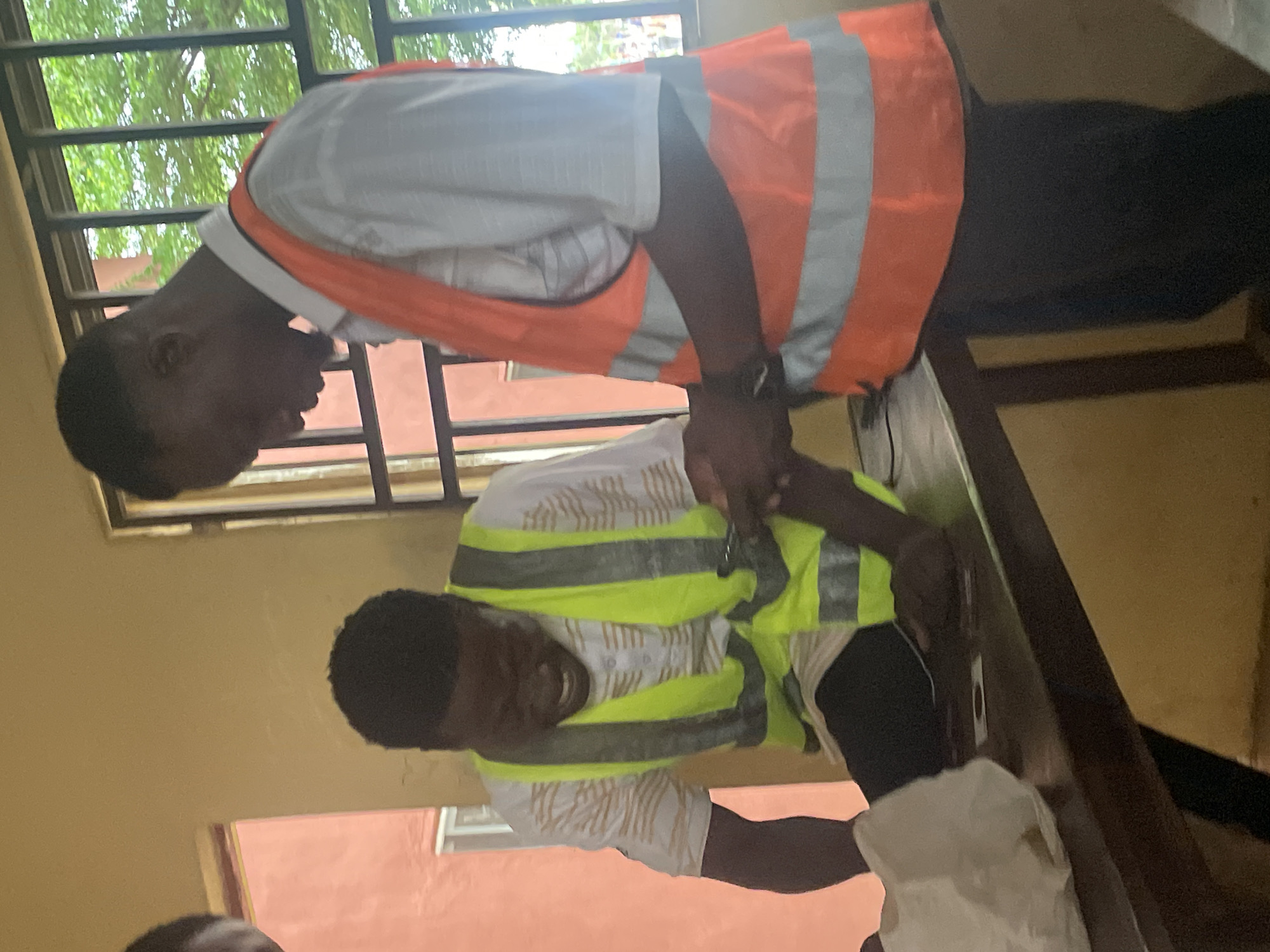 Two young men are standing at a wooden table in what appears to be a classroom or communal area in Lokoja, Nigeria. The man on the left is wearing a white shirt with a striped pattern and a bright yellow-green safety vest with reflective stripes. He is leaning forward, looking down at a mobile phone held in his hands, with his mouth open in what could be a laugh or exclamation. The man on the right is wearing a light-colored shirt and a bright orange safety vest with reflective stripes. He is also looking down at the phone, with his hands also engaged with the device. On the table in front of them are some small items, including what looks like a playing card and a white crumpled paper bag. In the background, there is a window with bars and green foliage visible outside, suggesting an outdoor area with trees. The walls of the room are a pale yellow color.
