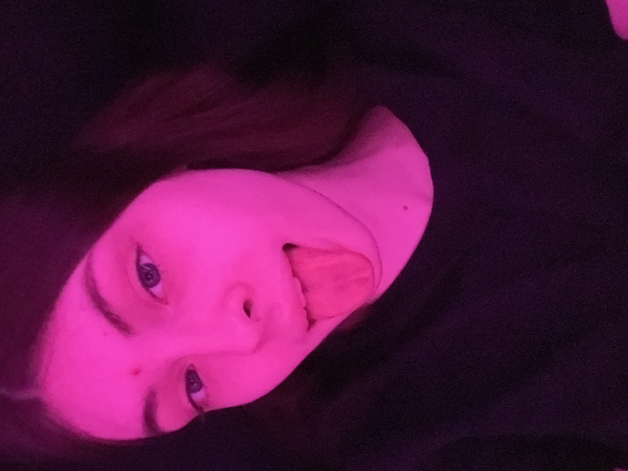 A photograph depicts a person's face illuminated in a pinkish-purple light. The person is sticking their tongue out. The background is dark, suggesting a low-light environment. The picture does not contain any elements related to the city of Saint Petersburg, Russia.
