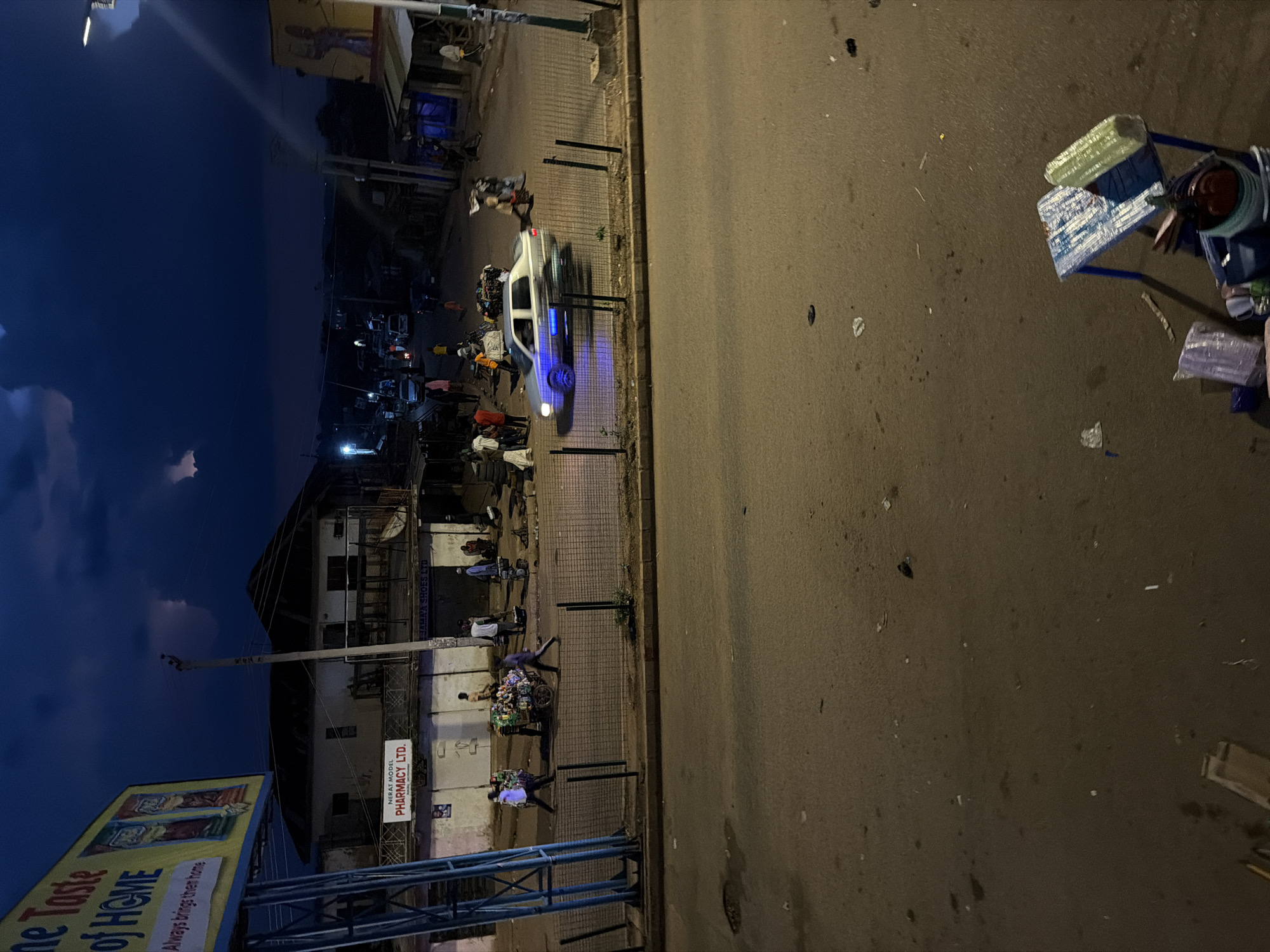 The image depicts a street scene at dusk in Bukuru, Nigeria. The sky is dark blue with visible clouds, and artificial lighting illuminates the area. In the foreground, a paved road shows scattered debris. A small, three-wheeled cart with blue metal legs and a white and blue patterned surface is visible on the right, carrying various items including what appears to be wrapped goods.
Further into the scene, a metal fence separates the road from a sidewalk area. On the sidewalk, several people are present. A light-colored sedan is driving on the road, with its headlights on and its undercarriage illuminated by blue lights. To the left of the road, a billboard advertising "Taste of HOME" is mounted on a blue metal frame.
Adjacent to the billboard is a building with a sign that reads "NERAT MODEL PHARMACY LTD." Several people are standing or walking near this building. Another building to the right has a sign that says "JAMAA SHOES LTD." In the background, more buildings and vehicles are visible, suggesting an urban or semi-urban environment. The overall impression is a busy street at the end of the day, with ongoing commercial and pedestrian activity.