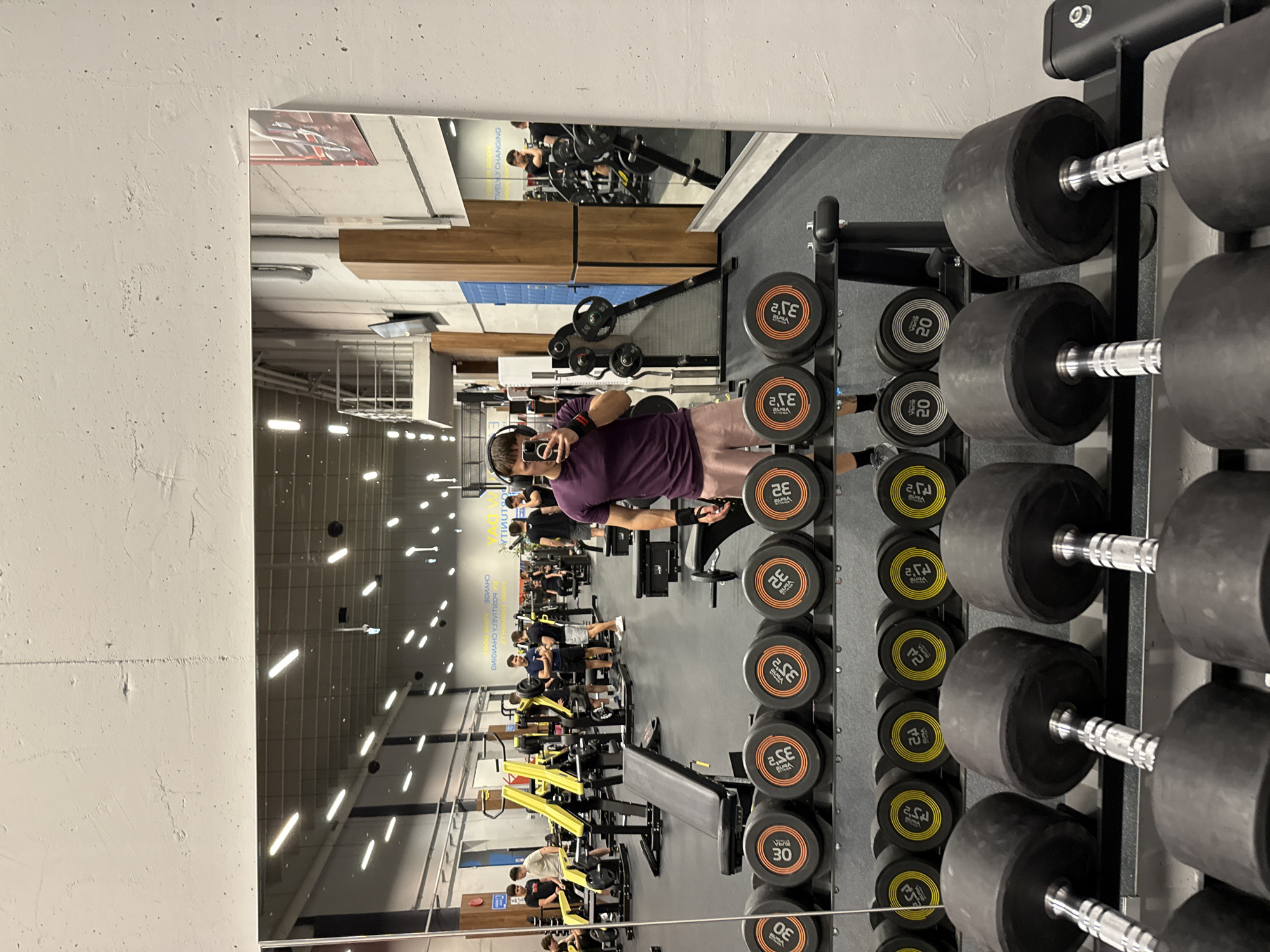 A gym interior is reflected in a mirror. The gym is long and filled with weightlifting equipment and people exercising. In the foreground, a rack of dumbbells stands to the right, with pairs ranging from small to large. A man, reflected in the mirror, stands in the middle ground taking a selfie with his phone. He wears a purple shirt and shorts and has headphones around his neck. The mirrored reflection reveals the depth of the gym, including various weight machines and benches occupied by people. Signage can be seen on the walls. Based on the visible signs, the gym is located in Sofia, Bulgaria.