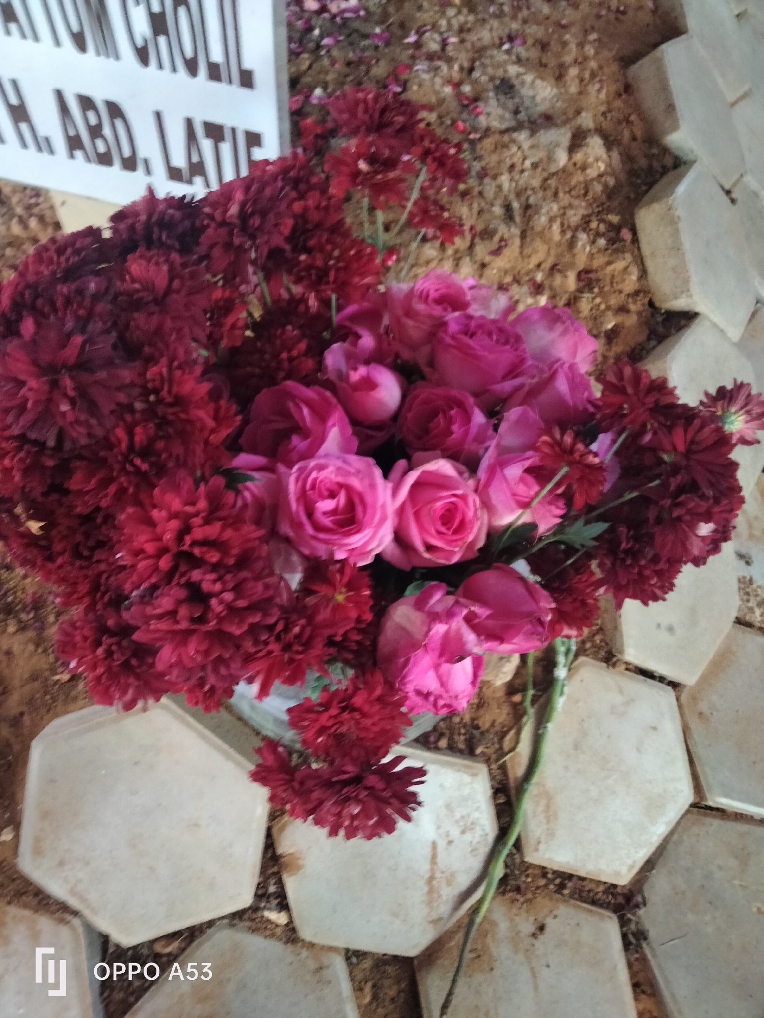 The image displays a bouquet of pink roses and dark red chrysanthemums placed on the ground, beside a tombstone. The tombstone, partially visible at the top left, has text inscribed on it, with visible portions reading "ATTUM CHOLIL" and "H. ABD. LATIE". The ground appears to be soil and is partly covered by hexagonal concrete tiles. The scene is outdoors, suggesting a cemetery or memorial site, possibly in the city of Badung, Indonesia, given the common use of such floral arrangements at gravesites in the region. The lighting suggests it is daytime. The bottom left corner of the image shows a watermark with the text "OPPO A53".