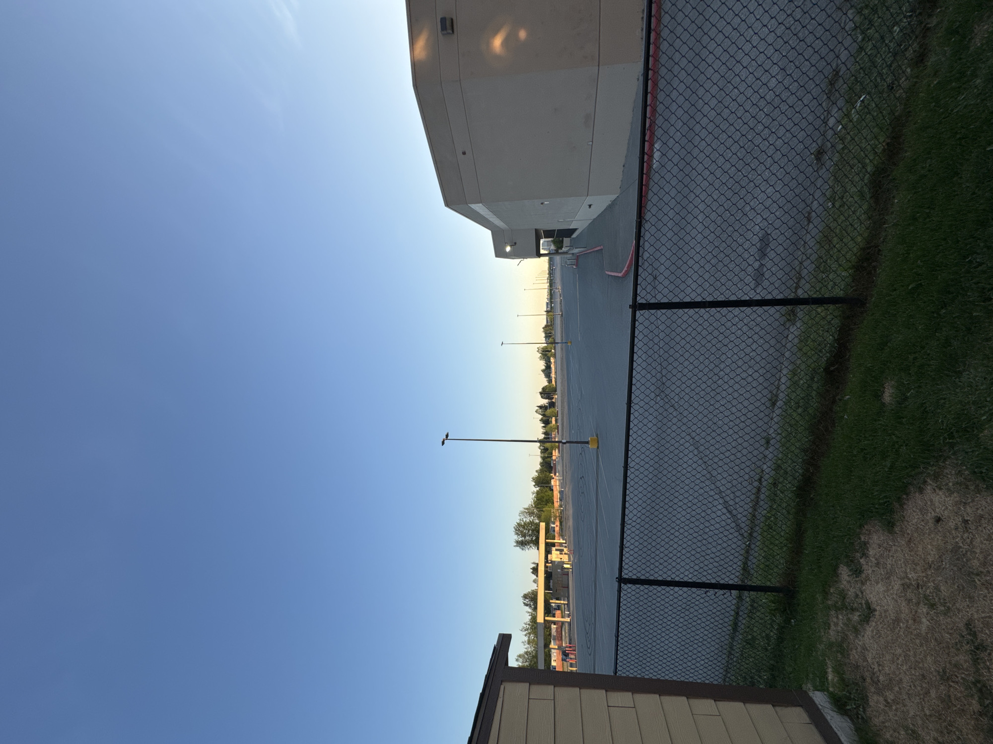 The image, when viewed upright, displays an outdoor scene encompassing a large commercial or industrial building, an extensive parking lot, and a chain-link fence under a clear sky. A substantial, light-colored building with a flat roof occupies the left side of the frame, showing architectural details consistent with a warehouse or large retail store. Its walls reflect warm light, suggesting a low sun angle.
A black chain-link fence stretches across the middle ground, acting as a boundary. To the right of the fence, there is a strip of green grass mixed with drier, lighter patches of earth. Beyond the fence, a wide, empty asphalt parking lot extends into the distance, dotted with numerous tall light poles. Some red painted lines are visible on the asphalt closer to the building. In the bottom right foreground, the corner of a shed-like structure, featuring light-colored horizontal siding and a dark roof, is partially visible.
The sky is a bright, clear blue with a hint of haziness or very thin clouds in the upper left. The soft, long shadows cast by the building and fence indicate either early morning or late afternoon/early evening. The parking lot appears deserted, with no vehicles or people present, contributing to a calm and quiet atmosphere. The scene suggests a commercial or industrial area within Anchorage, United States of America. There is no visible text in the image.