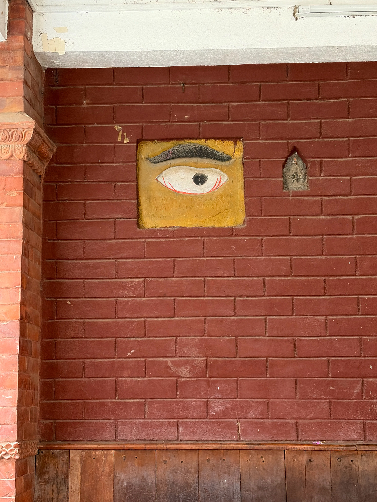 The image shows a close-up of a reddish-brown brick wall in Lalitpur, Nepal. On the wall, a square painting of an eye with a yellow background is mounted. To the right of the eye painting, a small, greyish idol is affixed to the wall. The wall is constructed of rectangular bricks laid in a stretcher bond pattern. A white painted ceiling ledge is visible at the top of the image, with peeling paint. A decorative brick pillar is visible on the left side of the image. The bottom portion of the image shows a wooden paneling.