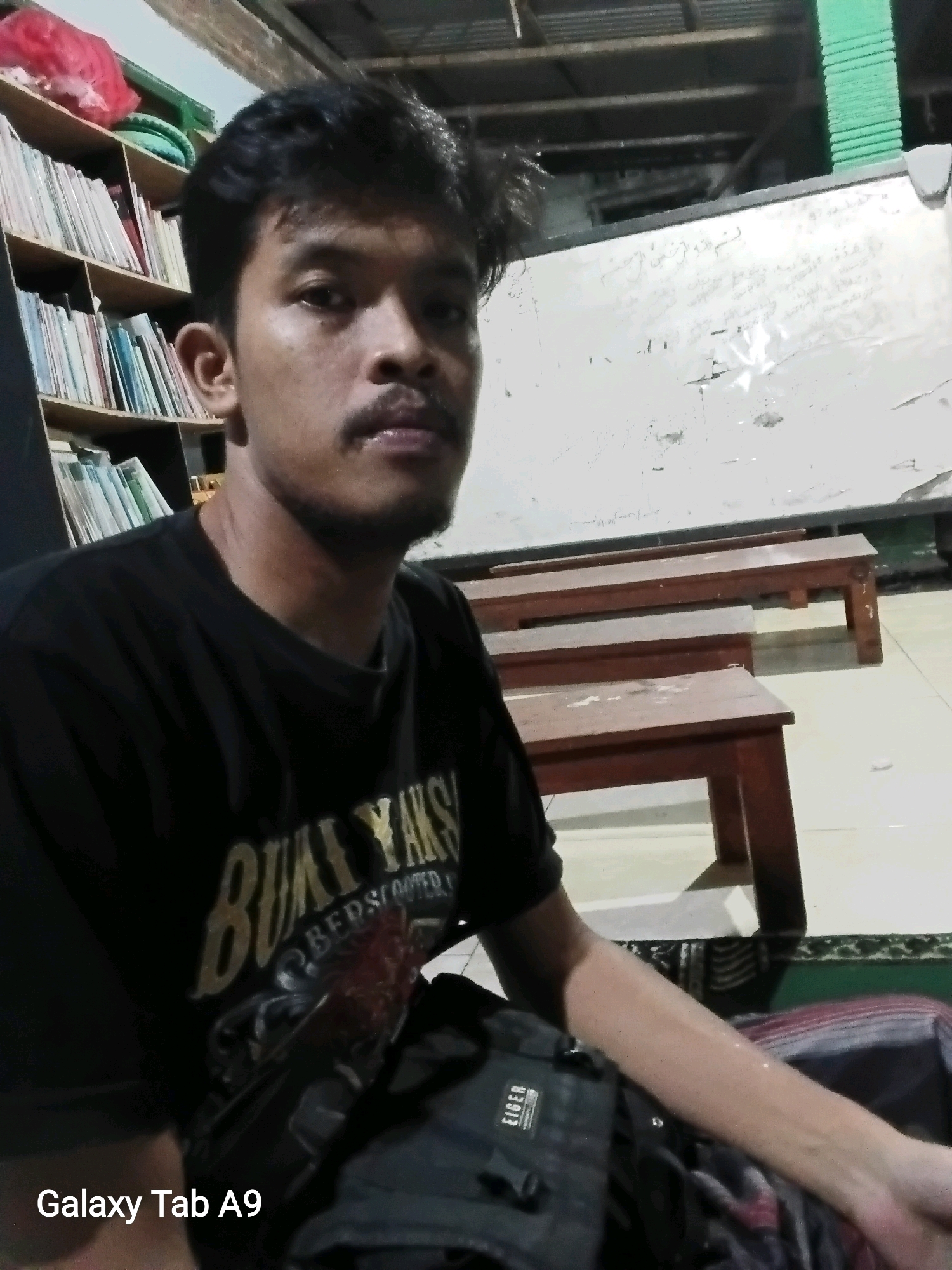 A young adult male with dark hair and a mustache is seated in front of a shelf filled with books. He is wearing a black t-shirt with a graphic print. To his right, there are two wooden benches arranged parallel to each other. Behind the benches, a whiteboard is visible, covered in handwritten text. The overall environment suggests an indoor setting, possibly a classroom or library. The lighting is dim, casting shadows on the scene. The bottom left corner of the image displays the text "Galaxy Tab A9". The image has a timestamp suggesting it was taken on an unspecified date at an unspecified time. The location is identified as Trenggalek, Indonesia.