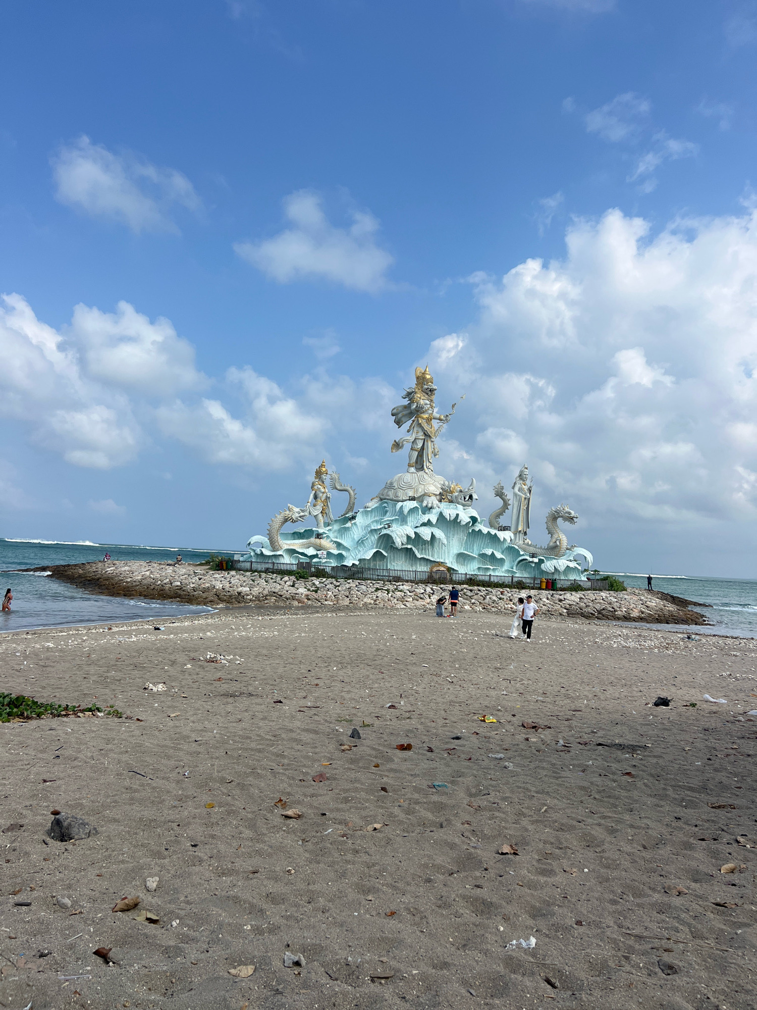 The image depicts the GWK Cultural Park's Lord Vishnu statue in Kuta, Indonesia, on a sunny day with scattered clouds. The statue is a prominent feature, with a large figure of Vishnu at the apex, surrounded by smaller statues and dragon-like sculptures, all situated on a rocky embankment. The sea stretches out behind the monument, with waves gently rolling towards the shore. In the foreground, a sandy beach is visible, littered with debris, including leaves, stones, and various pieces of trash. A few people are present on the beach and near the statue, some walking and others observing the monument. The overall scene captures a blend of natural beauty and cultural artistry, with the beach serving as a viewpoint for the grand statue.