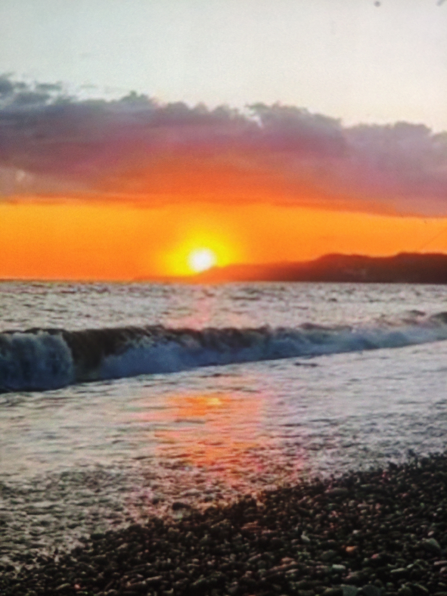 The image depicts a sunset over a body of water, likely the Black Sea, with a pebble beach in the foreground. The sky is filled with vibrant orange and pink hues, with a bright sun visible near the horizon. Dark clouds are present in the upper part of the sky. Waves are crashing on the shore, and the water appears to be a deep blue. The pebble beach is covered in dark-colored stones. There are no people or specific structures visible in the image. The location is identified as Armavir, Russia, though Armavir itself is inland and not on the coast.