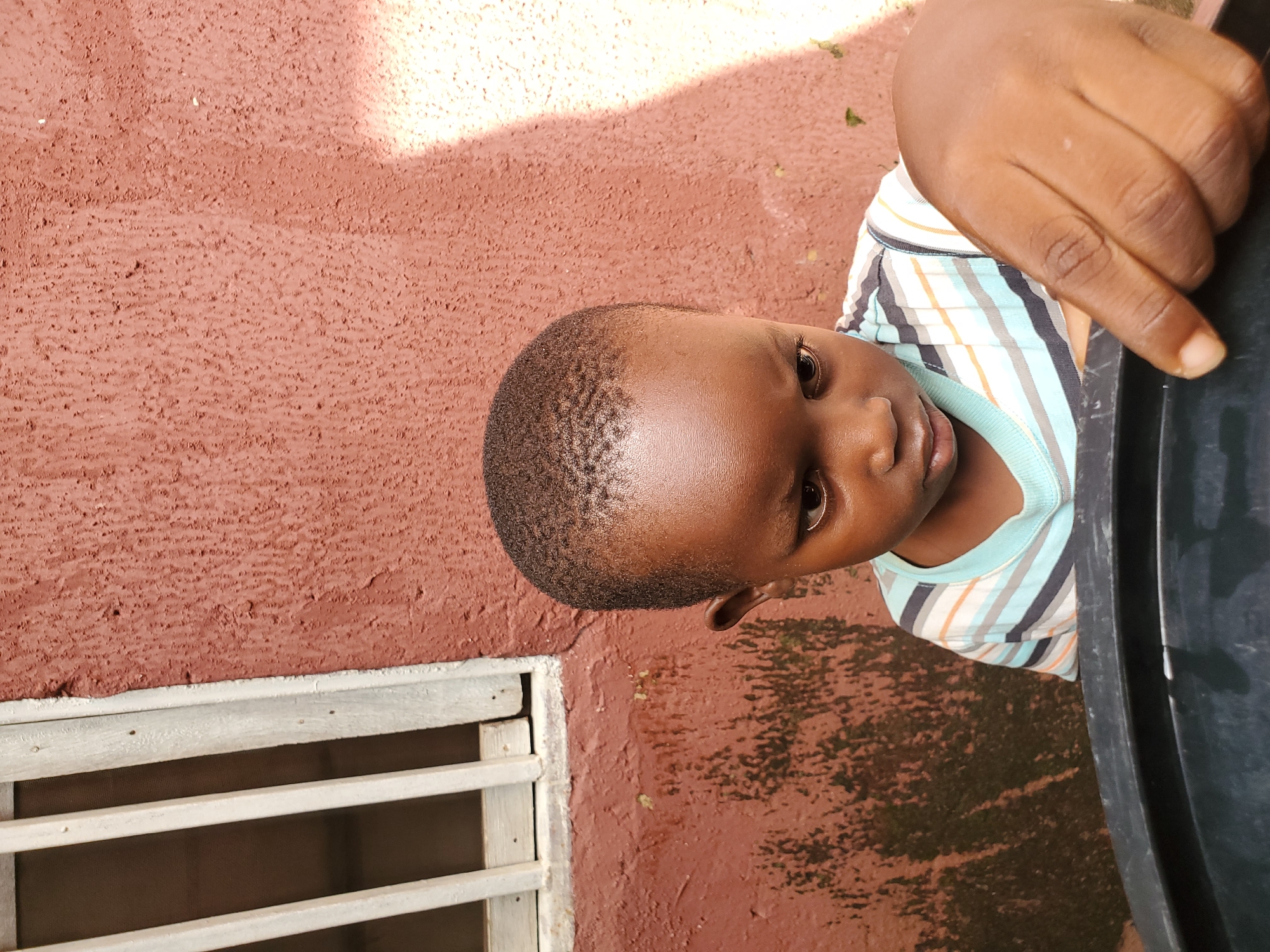 The image shows a close-up, side-profile view of a young Black child looking towards the camera. The child has short, dark hair and is wearing a striped shirt with blue, white, orange, and black stripes. A dark object, possibly a large container, is visible to the right of the child. The child's left hand is also visible, resting on or near the container.
The background consists of a textured, reddish-brown wall, with a patch of sunlight illuminating the upper right portion of the image. A window with horizontal wooden slats is visible in the lower left quadrant, suggesting an outdoor or semi-outdoor setting. The ground appears to be wet or stained in patches.
The lighting suggests it is daytime, possibly sunny, given the bright highlight on the wall. The overall impression is one of a casual, everyday moment captured outdoors. No text is visible in the image.