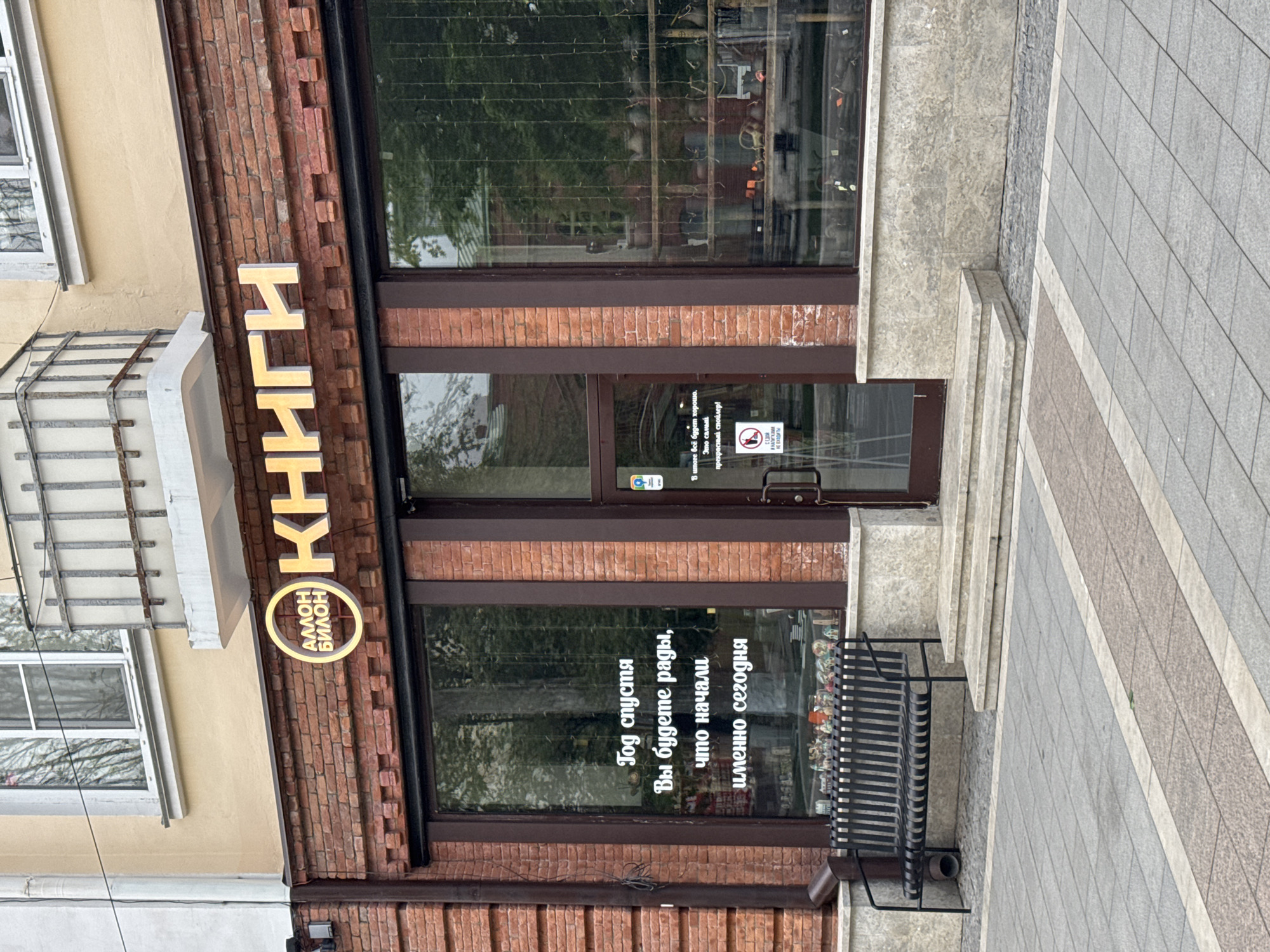 The image depicts the storefront of a bookstore in Akhieli, Georgia. The sign reads "Books Amon Bilon" vertically along the red brick facade of the building. The windows have a quote on them "A year from now you'll be glad you started today." A black bench is in front of the window. The entrance has a dark-framed glass door with signage including "No smoking". The storefront is adjacent to a sidewalk. The building is several stories high.