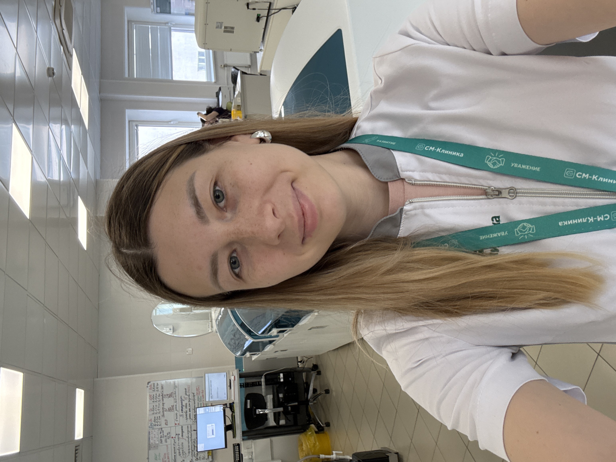 A medium shot, selfie-style photograph captures a woman with blonde hair and blue eyes in a laboratory setting. She is wearing a white lab coat over a pink shirt and a teal lanyard with the text "СМ-КЛИНИКА" (SM-CLINICA) and iconography. She has freckles on her nose and cheeks and is smiling at the camera.
The background reveals a clinical environment with medical equipment. To the left, a rolling cart holds two computer monitors and a black office chair. A yellow biohazard bin is visible on the floor. In the mid-ground, a long, white medical analyzer with a teal-colored top is prominently displayed. Further back, windows with blinds are visible, and another piece of medical equipment, possibly a large cabinet or another analyzer, is to the right. A whiteboard with handwritten text is mounted on the wall. The floor is tiled with a grid pattern. The overall impression is that of a modern, well-equipped medical facility. The presence of the "СМ-КЛИНИКА" branding on the lanyard strongly suggests the location is a branch of the SM-CLINICA medical network, which has multiple facilities in Moscow, Russia.