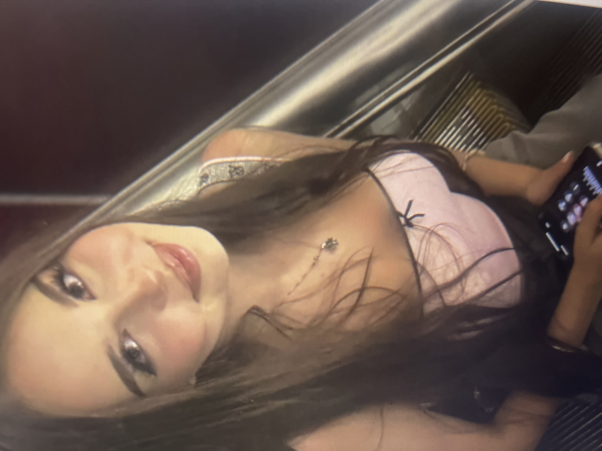 A young woman with long, dark hair is captured in a close-up, upward-angled shot. She has fair skin, prominent eyelashes, and is wearing light-colored makeup. Her lips are slightly parted, and she appears to be smiling. She is dressed in a light pink top with black trim and a small black bow detail at the center. A delicate silver necklace with a pendant rests on her collarbone. Her left hand is visible, holding a smartphone. The background is a blurred, metallic, ribbed surface, suggesting an escalator or similar urban structure. The lighting is dim, creating a soft, diffused effect. There is no discernible connection to the city of Qarağandı city administration, Kazakhstan.