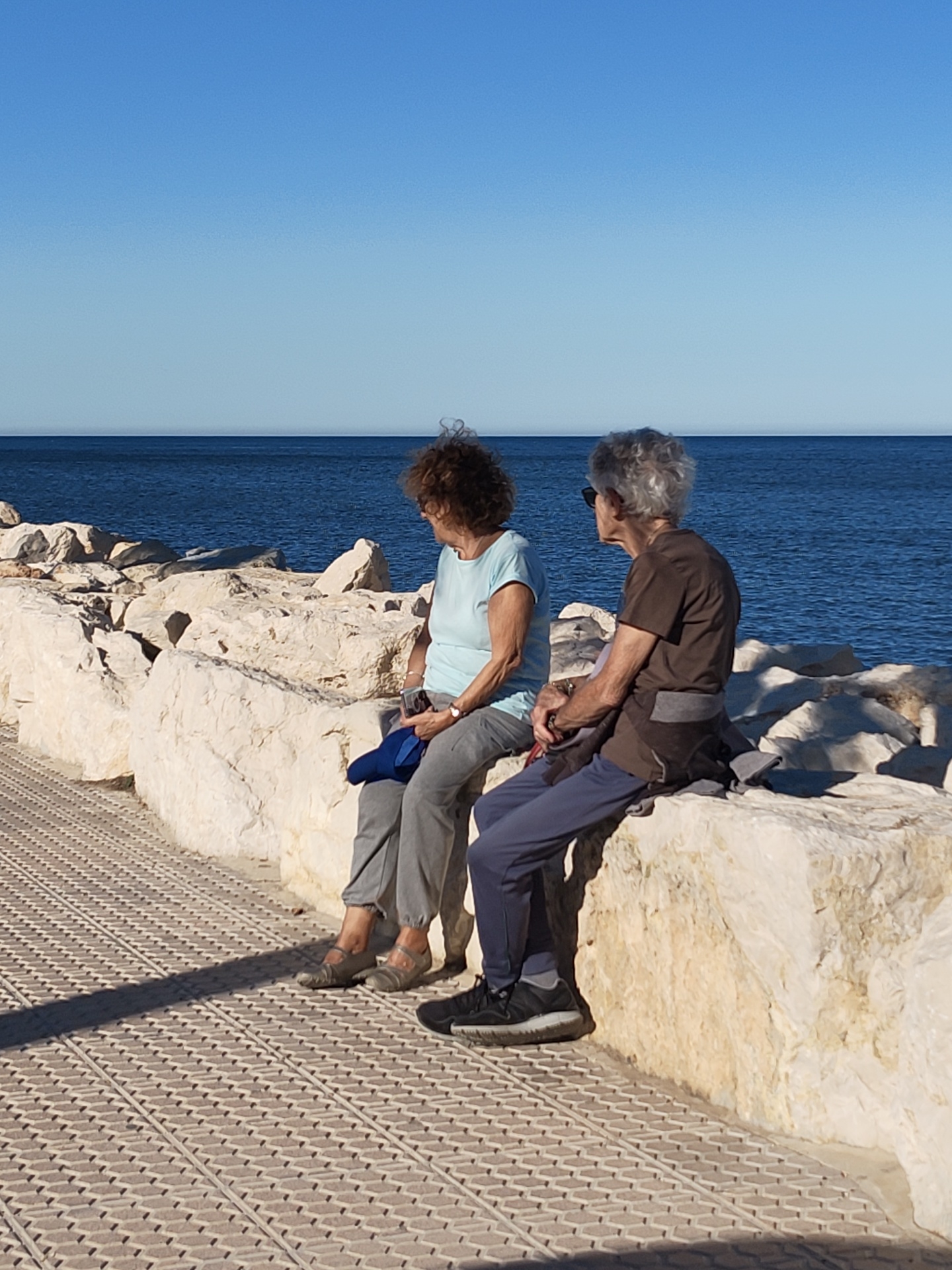 This outdoor photograph captures two older women seated on large, light-colored rocks by the sea, under a clear, bright blue sky, likely on a sunny day in Dénia, Spain. The image is taken from a slightly elevated perspective, looking towards the sea.
The woman on the left has brown, curly hair and is dressed in a light blue t-shirt, grey pants, and light-colored flat shoes. She holds a dark blue item, possibly a hat or folded garment, and a small dark object in her lap. A watch is visible on her left wrist. The woman on the right has grey, curly hair and is wearing sunglasses, a brown t-shirt, dark blue pants, and black sneakers. A dark grey garment is tied around her waist. Both women are facing to the right of the frame, appearing to gaze out at the calm, deep blue sea that meets the horizon under the cloudless sky.
In the foreground, a light-colored paved walkway with a distinctive grid or diamond pattern stretches across the bottom left of the frame. Strong sunlight casts sharp shadows from the women and the rocks onto this path. The rocks form a natural barrier or breakwater between the walkway and the sea. The scene exudes a sense of tranquility and leisure, suggesting a relaxed moment enjoying the coastal view. No text is visible in the image.
