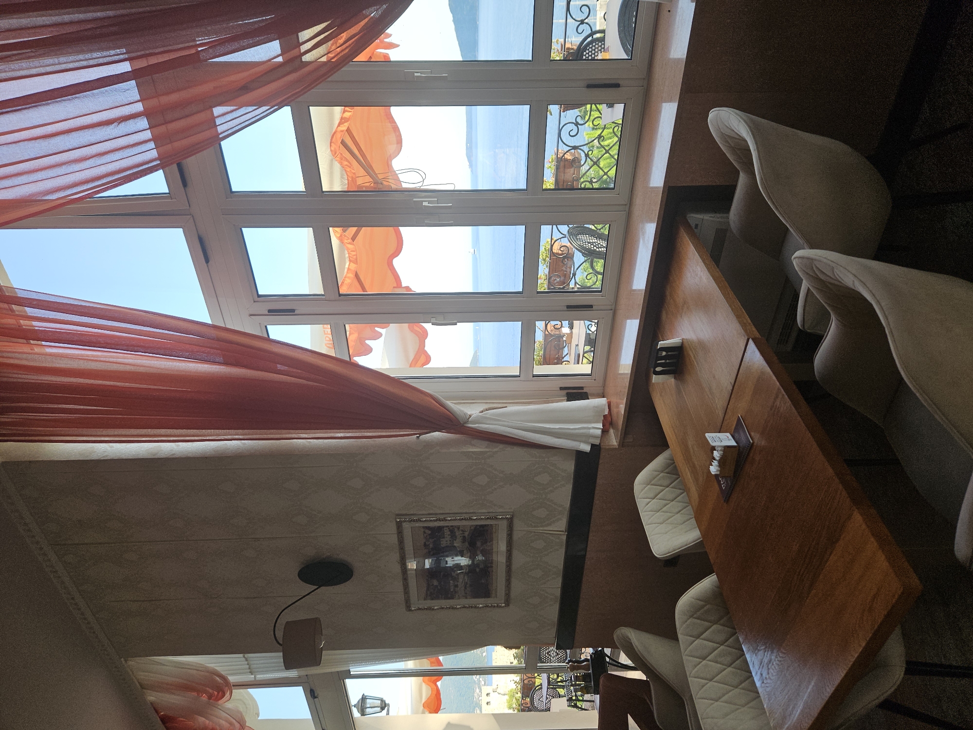 This media file shows a dining area with a view of the sea, likely in Herceg Novi, Montenegro. The scene is set indoors, in what appears to be a restaurant or cafe. Large windows dominate the scene, offering a panoramic view of a bright blue sea under a clear sky. Sheer, orange-red curtains are drawn back, framing the windows and adding a touch of color to the interior.
Inside, a polished wooden table is set for dining, with white quilted chairs arranged around it. On the table, a small stack of napkins with cutlery is visible, along with a small card holder. The interior walls are decorated with a subtle patterned wallpaper. A framed picture hangs on one wall, depicting what looks like a European town.
The bright sunlight streaming through the windows suggests it is daytime, possibly late morning or early afternoon. The calm sea and clear sky indicate pleasant weather conditions. Outside the windows, glimpses of balconies with wrought-iron railings and potted plants suggest an outdoor seating area or promenade. In the distance, a sailboat can be seen on the water, adding to the serene coastal atmosphere. The overall mood of the image is tranquil and inviting, highlighting the picturesque view and comfortable setting.
Visible text includes "SPEZI" and "ALIA" on a menu or placemat on the table.