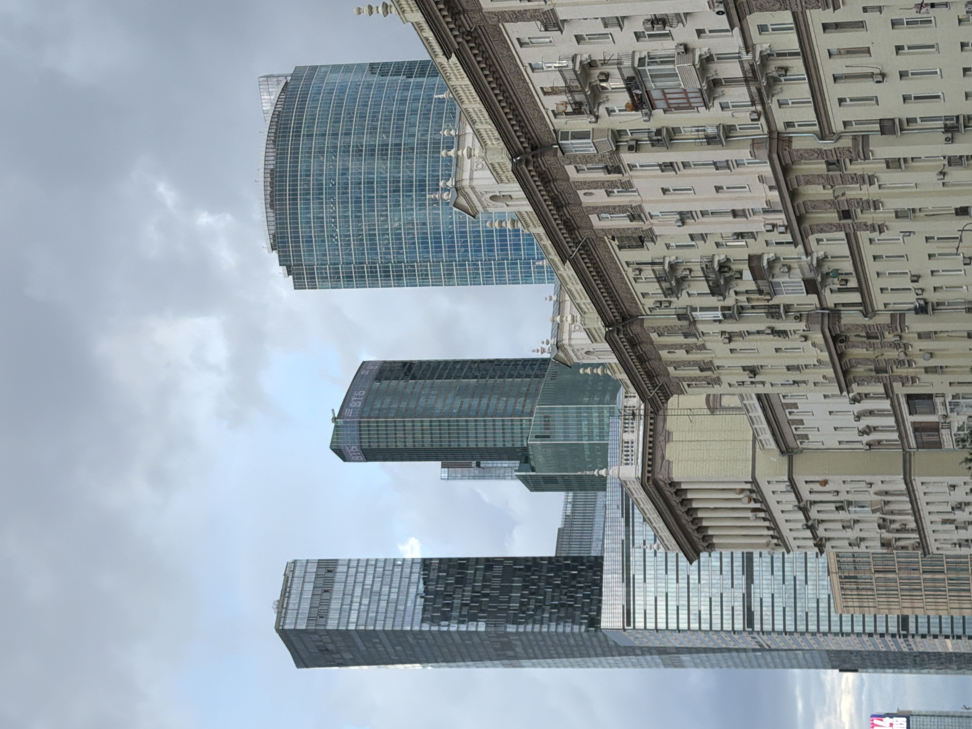 The image depicts a view of Moscow's Moskva-City business district under an overcast sky. In the foreground, an older, light-colored building with ornate architectural details and balconies spans the width of the frame. Behind this building, several modern skyscrapers rise into the sky. The tallest and most prominent skyscraper on the left is a dark, rectangular glass tower with a segmented facade. To its right is a teal-colored skyscraper with the "ВТБ" logo visible. Further to the right is a rounded, blue glass skyscraper. The sky is filled with heavy, gray clouds, suggesting a cool or rainy day. The scene is devoid of any people or visible activity, focusing solely on the architecture and urban landscape.