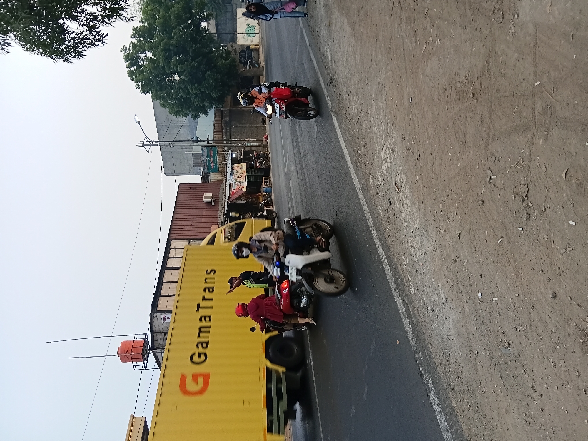 This is a street-level, full-shot, eye-level photograph taken in Bekasi, Indonesia, under an overcast sky. The scene captures a busy intersection with several motorcycles and a large yellow truck.
In the foreground, the asphalt road is visible with a white lane divider. The right side of the road is a dirt and gravel shoulder with scattered debris.
Dominating the left side of the image is a bright yellow shipping container on a truck, branded with "GamaTrans." A portion of the truck's cab and wheels are visible.
In the mid-ground, several motorcycles are present. A red motorcycle with a rider wearing a red helmet and jacket is in the left lane, stopped. To the right of this is a white and blue motorcycle with a rider wearing a light-colored jacket and a mask, also stopped. Behind these two, a traffic officer in a yellow and black vest and dark pants is directing traffic, with their right arm raised. Further to the right, a red motorcycle is moving, with two people on it, one wearing an orange vest.
In the background, there are buildings, including a red corrugated metal structure and a grey concrete building. A water tower with an orange tank sits on top of a building on the far left. Trees, including a large leafy tree on the right, are present. A street lamp is visible on a pole in the mid-background. Two women are walking on the right side of the road in the background, one wearing a blue hijab and carrying a pink bag.
