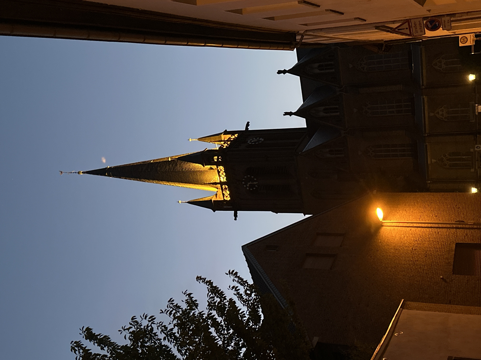 The image shows a view of the historic Protestant church, Lutherkirche, in Krefeld, Germany, at dusk. The church's illuminated spires and Gothic architecture dominate the center of the frame, with the warm glow of artificial lights contrasting against the darkening blue sky. To the left, the silhouette of a brick building and a tree are visible, partially obscuring another building. To the right, the side of a building with a distinctive cream-colored facade is seen, with a street sign indicating "Straßenschäden" (road damage) and other traffic symbols. A streetlamp casts an orange glow on the brick wall of the building on the left. The scene depicts a quiet evening in an urban environment, highlighting the architectural heritage of Krefeld.