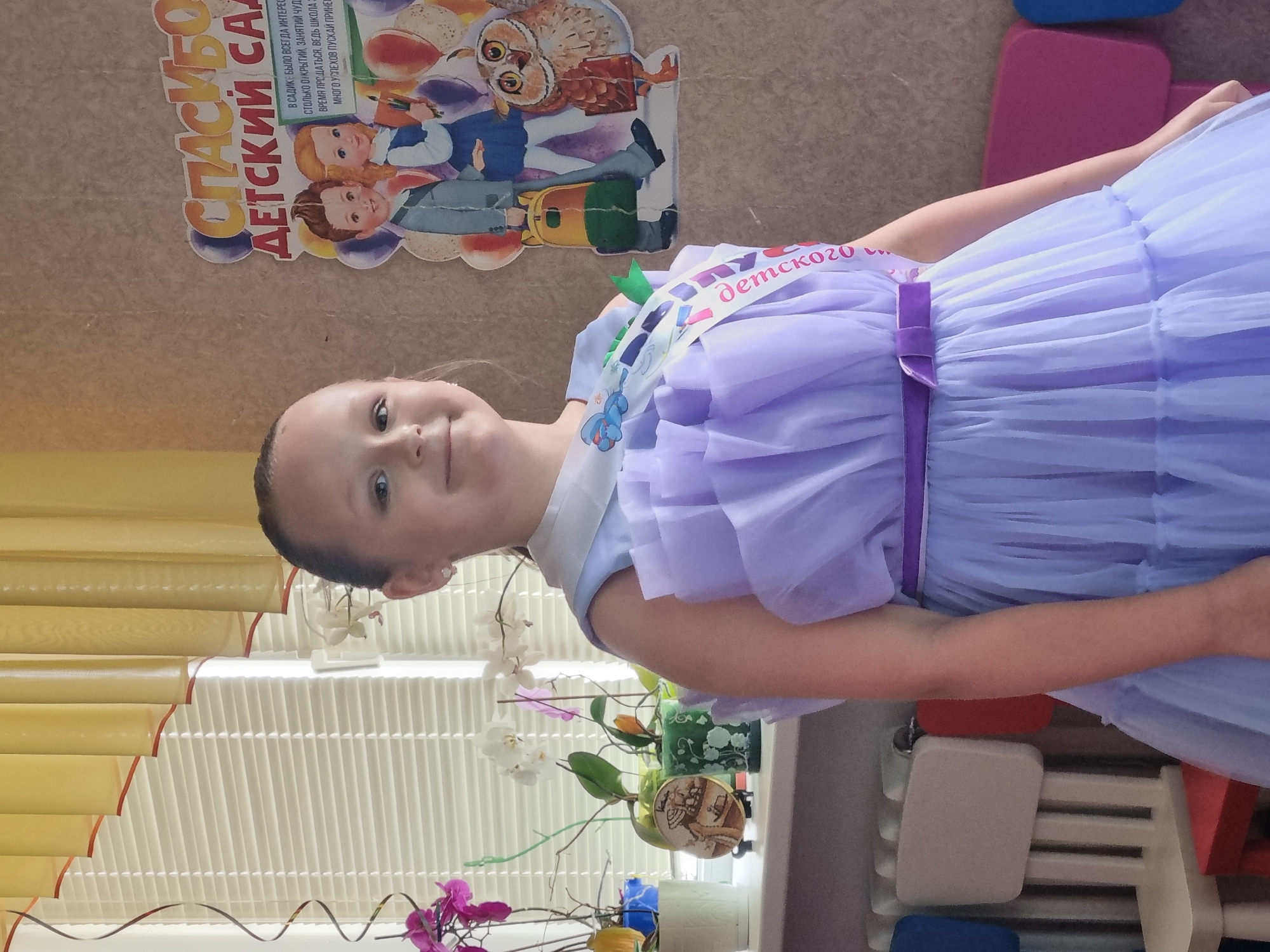 The image shows a young girl in a purple dress, wearing a sash that reads "Детского" (Kindergarten). She appears to be at a graduation or end-of-year celebration for kindergarten. The background includes a colorful poster depicting children and an owl, with text that reads "СПАСИБО ДЕТСКИЙ САД" (Thank you, Kindergarten). There are also blinds in the background and some plants. The girl is smiling, suggesting a happy and celebratory mood. The context suggests this is in Buzuluk, Russia.