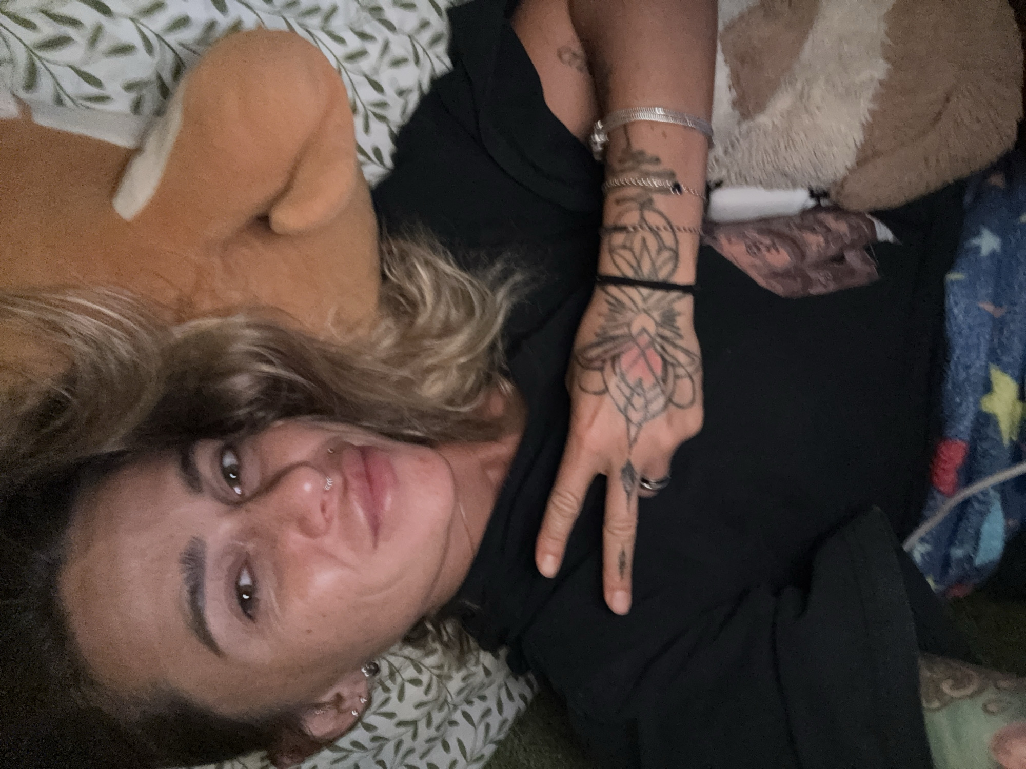 The image shows a person lying down, possibly in a bed, given the presence of pillows and blankets. The person has blonde hair and is looking at the camera. They are wearing a black top and have several tattoos on their arm and hand, including one that resembles a mandala. They are also wearing jewelry, such as bracelets and rings, and have piercings on their nose and ear. Next to the person is a stuffed animal, a portion of which is visible. The background appears to be a bed with a patterned sheet and a colorful blanket. The lighting suggests it is daytime. The person is making a peace sign with their hand. The setting does not provide strong cues for the specific location within Thep Krasatti, Thailand, but the indoor, relaxed atmosphere is evident.