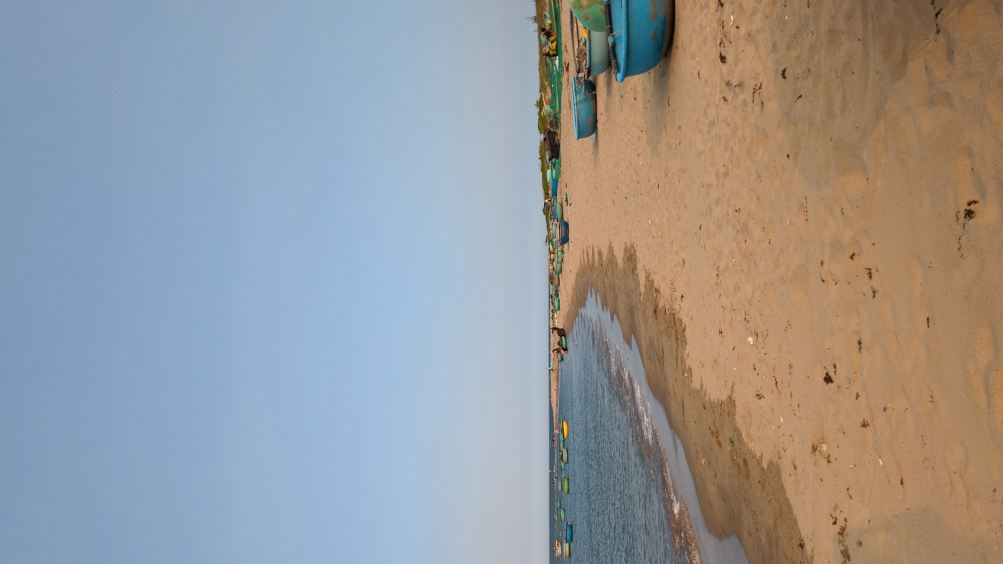 This landscape photograph captures a beach scene in Phan Rang – Tháp Chàm, Vietnam. The beach stretches along the coastline with sand meeting the sea. A row of blue boats rests on the sand near the water's edge, extending into the distance. The ocean waves gently approach the shore. The sky is a clear, pale blue. A few small figures can be seen on the beach.