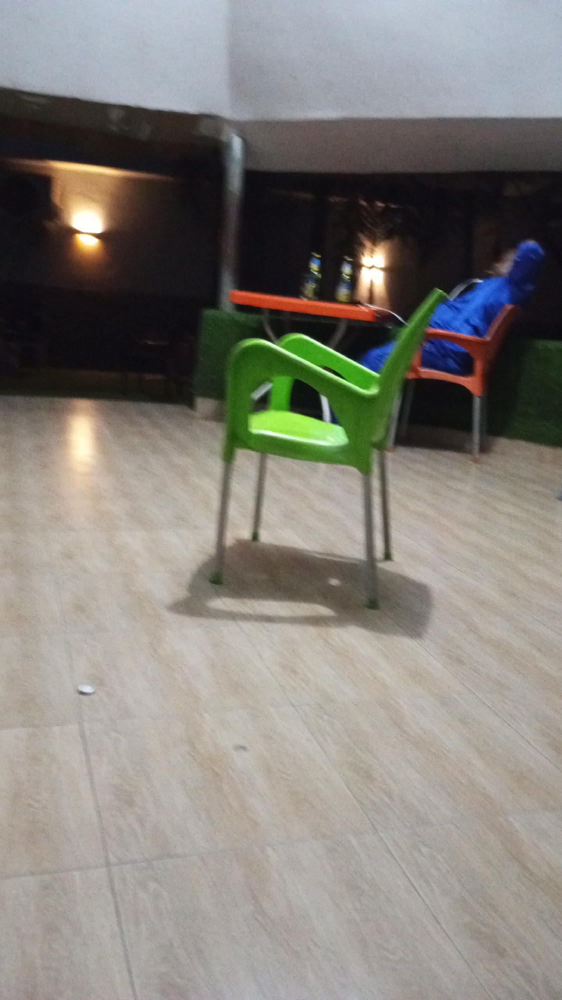 The image shows an outdoor seating area in Adankolo, Nigeria. The foreground features a bright green plastic chair on a tiled floor. In the background, there is an orange table with two bottles on it, and a person in a blue garment is sitting on an orange chair. A light is visible in the background and green plant decor is present behind the chairs. The lighting suggests it is evening or night.