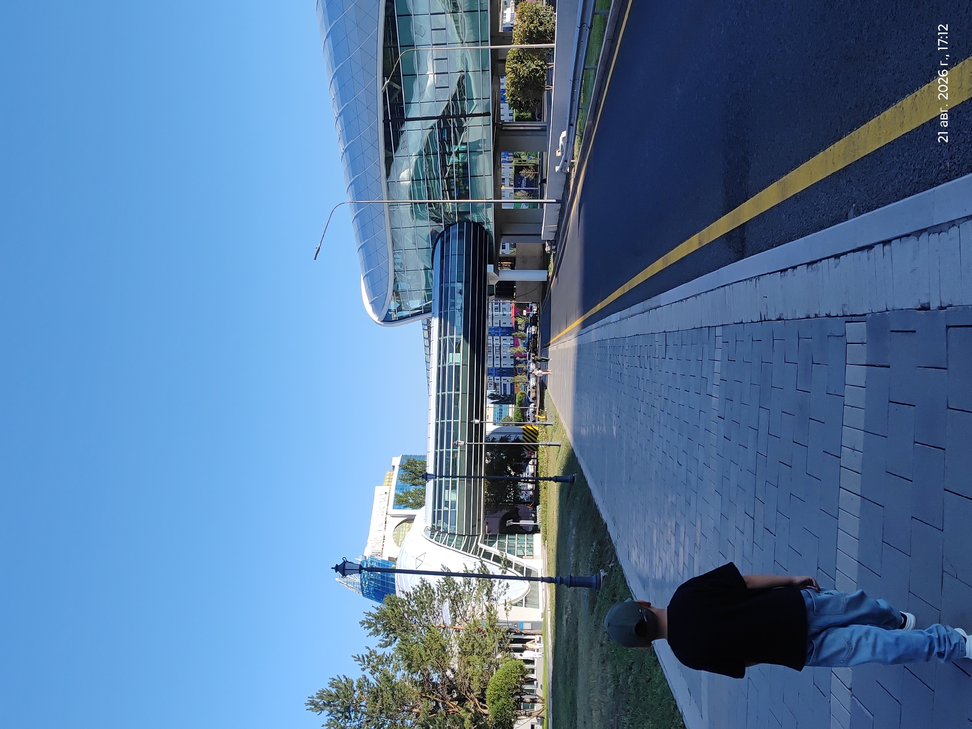 The image depicts a young boy in a black t-shirt and blue jeans, wearing a green cap, walking away from the camera on a paved sidewalk. The sidewalk is bordered by a grassy area on the left and a road with a yellow line on the right. In the background, there is a modern architectural complex with glass facades and elevated walkways. The sky is clear and blue, suggesting a sunny day. The timestamp at the bottom right corner indicates "21 Aug, 2026, 17:12". The architectural style and the presence of the building complex, which appears to be the Nazarbayev University or a similar modern facility, strongly suggest the location is Astana, Kazakhstan.