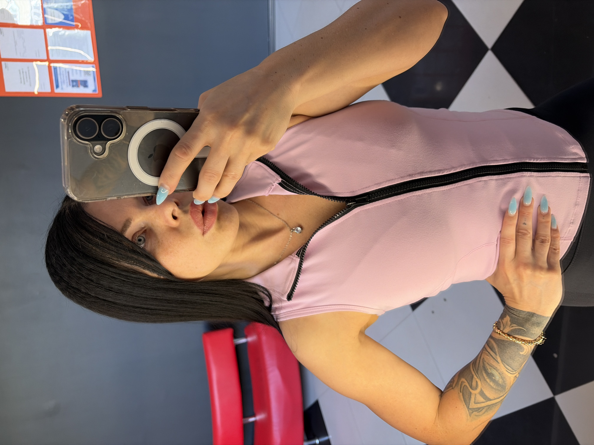 The media file shows a woman taking a selfie in a room. She has long dark hair and is wearing a light pink zippered top and black leggings. Her nails are painted a light blue color. She is wearing a necklace with a small pendant. She has a tattoo on her left arm and is wearing a bracelet. In the background, there is a red chair and a tiled floor with a black and white checkerboard pattern. There are also some documents displayed on a board on the wall. The lighting suggests it could be daytime.