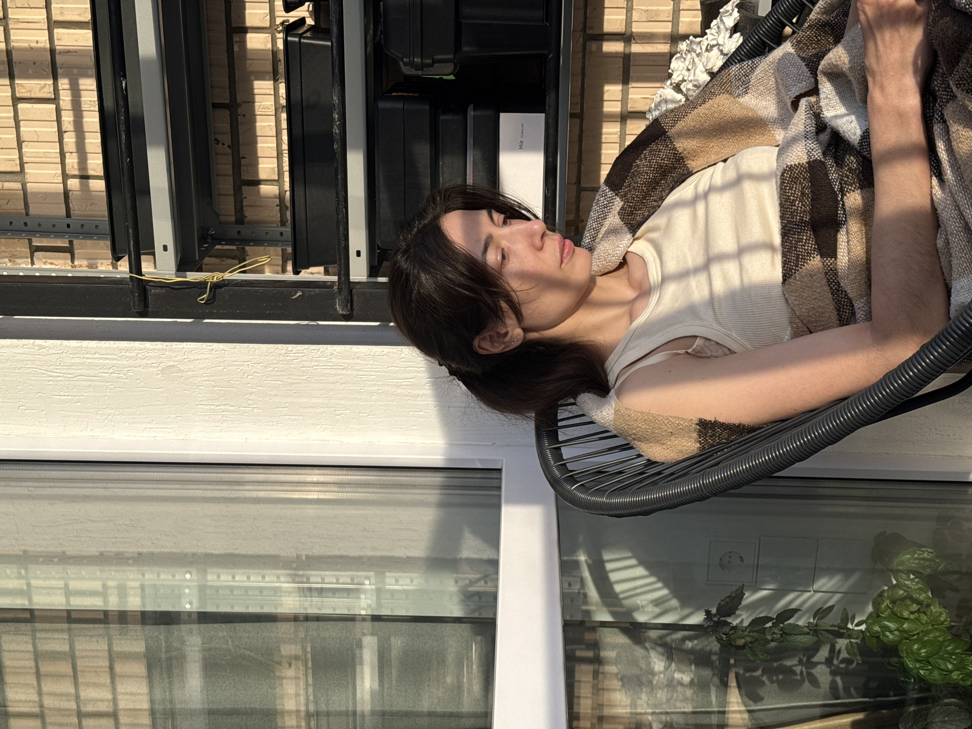 A person with dark hair is reclined in a gray circular chair, wrapped in a brown and white plaid blanket. The individual is fair-skinned and wearing a light-colored tank top. Their eyes are closed, suggesting they may be resting or sleeping.
Behind the person, a window with white framing shows reflections of outdoor elements. A plant with green leaves is visible through the window, and further back, there's a brick or stone wall.
The composition of the image is such that the person and chair are positioned to the right side, with the window occupying the lower left portion. A set of black storage containers or cases are stacked on a shelf to the upper left, secured by a yellow twist tie.
The image lacks specific details that would reveal its location in Perm, Russia; the scene appears to be indoors, near a window that may offer a view of the external environment.