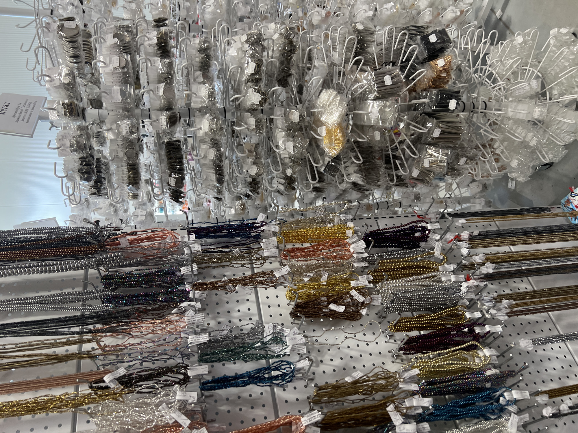 The image shows a display of beads and metal findings inside a store. The lower half of the image presents rows of colorful bead strands hung on hooks attached to a white, perforated pegboard. The strands are various colors, including gold, silver, bronze, and shades of blue, green, and purple. Above the beaded strands, there are wire racks holding packets of metal findings and beads in clear plastic packaging. The packets have small white tags attached. A sign with Cyrillic text labeled "Metal" is visible on the left side of the image. The store's lighting is bright, and the overall setting is organized and commercial. The location is a store selling craft and jewelry-making supplies in the city of Burgas, Bulgaria.