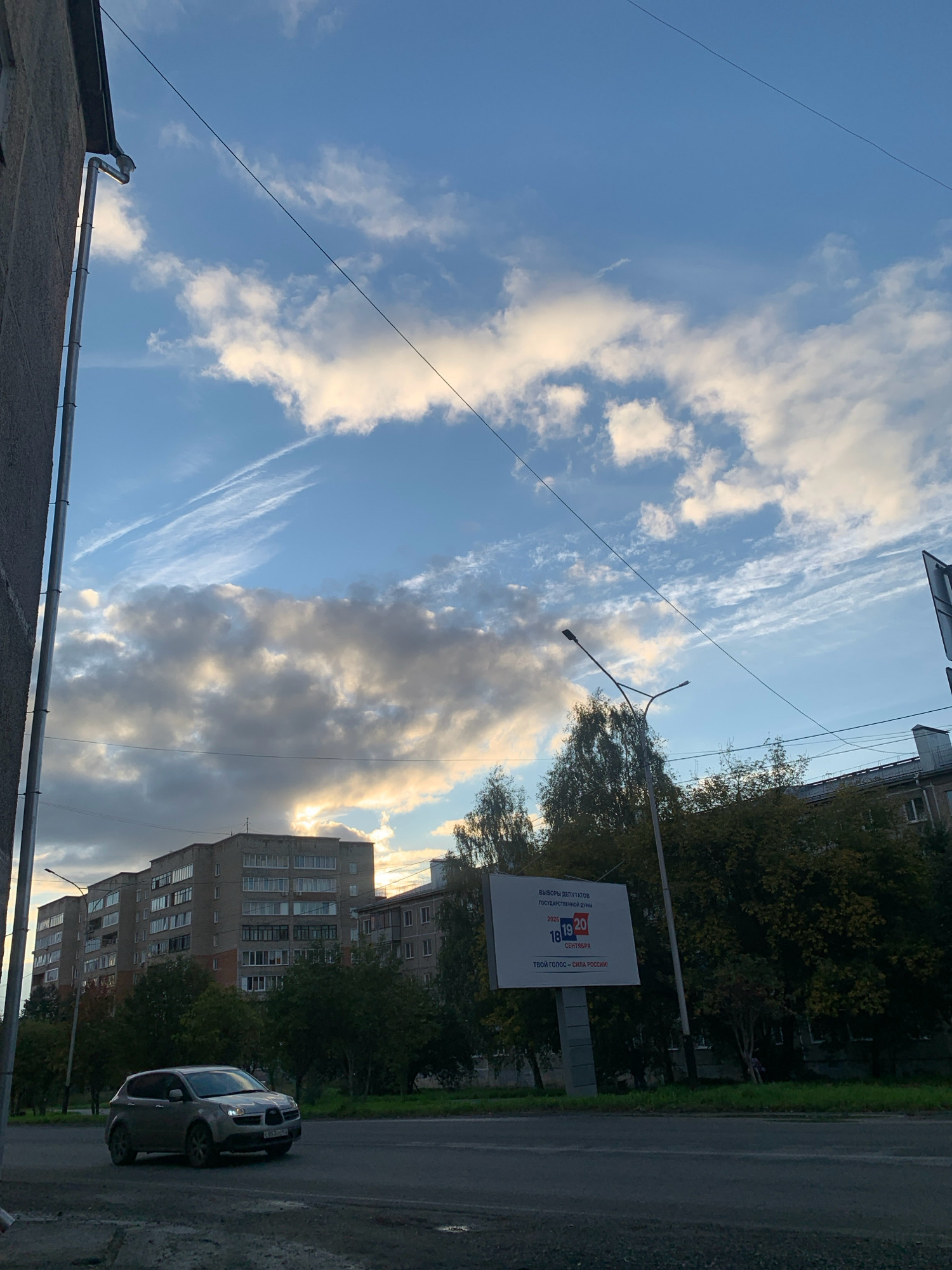 This image captures an urban scene in Prokopyevsk, Russia, under a dynamic sky. The perspective is from a street level, looking towards a cluster of apartment buildings and trees. In the foreground, a grey car is driving on a wet asphalt road. Overhead, a large billboard displays text and numbers, with "18 19 20 СЕНТЯБРЯ" prominent, suggesting an election date. The sky above is a mix of blue and grey clouds, with sunlight breaking through in some areas. A streetlight is visible, and power lines crisscross the sky. The architecture of the buildings suggests a post-Soviet era construction style. The overall impression is of an ordinary day in a Russian city, with a hint of political messaging from the billboard.