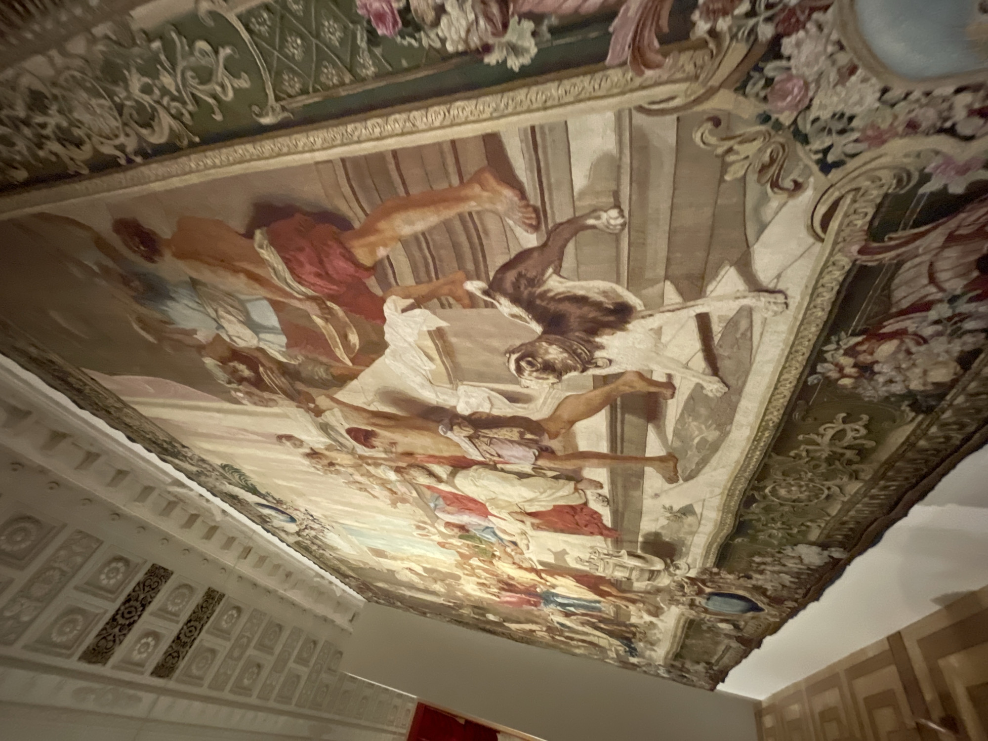 This image shows a large, ornate tapestry hanging on a wall in a room in the Hermitage Museum in Saint Petersburg, Russia. The tapestry depicts a scene with many figures, possibly from a historical or mythological event, with a dog in the foreground. The room has a high, decorated ceiling and what appears to be a parquet floor. The tapestry itself is richly detailed with many figures in classical attire, vibrant colors, and intricate borders. The lighting in the room is somewhat dim, creating a dramatic effect on the tapestry.