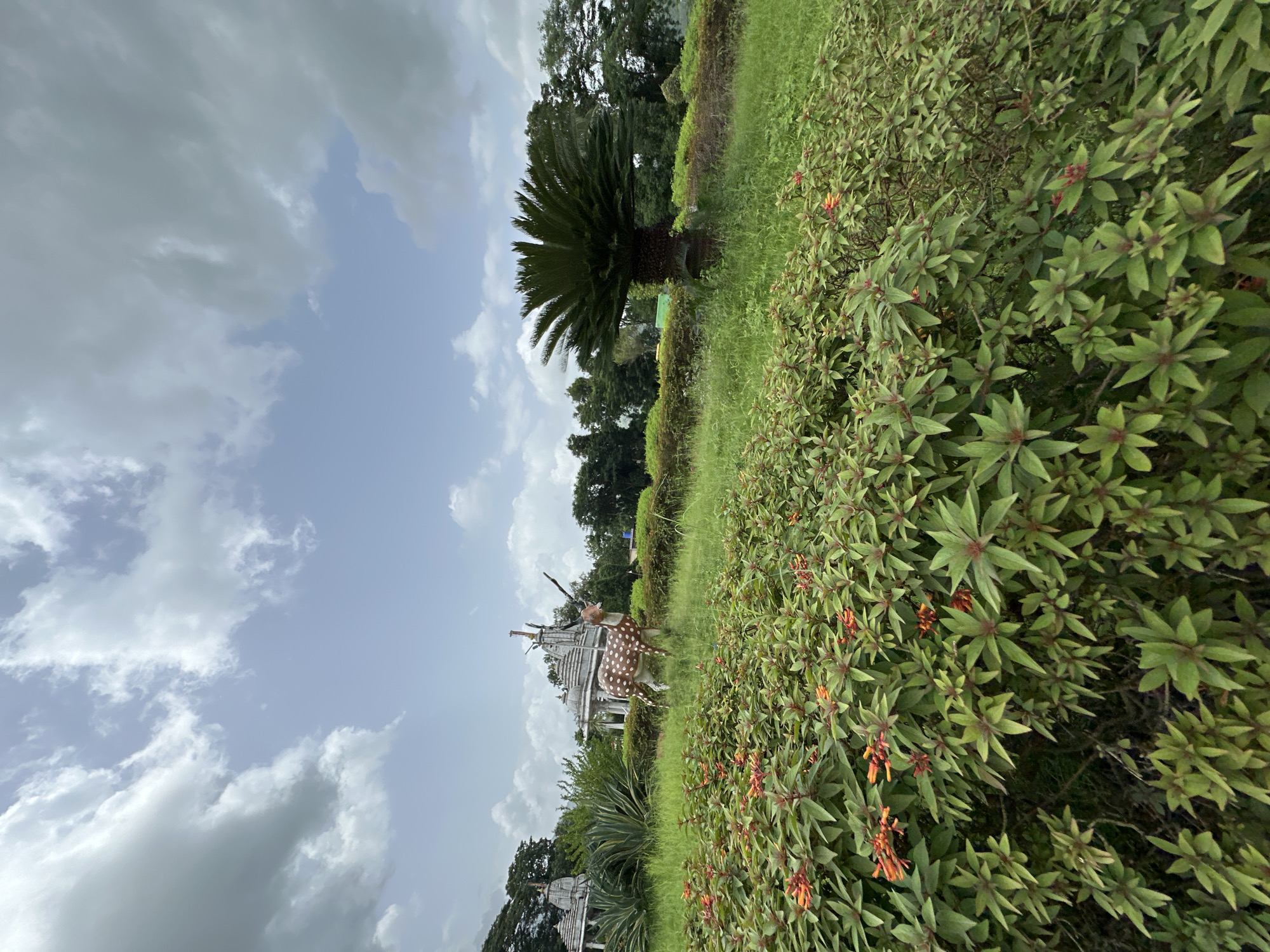 The image depicts a serene outdoor scene, likely within a park or garden, in Shahapur, India. In the foreground, lush green bushes with small orange flowers are in bloom. Behind these, a grassy area is dotted with more greenery, including a tall palm-like plant and a decorative statue of a spotted deer. In the mid-ground, a white gazebo with intricate architectural details stands amidst the foliage, and further back, more white structures, possibly temples, are visible with trees forming a backdrop. The sky above is partly cloudy, with a mix of white and grey clouds against a pale blue expanse. The overall atmosphere suggests a tranquil and well-maintained public space.