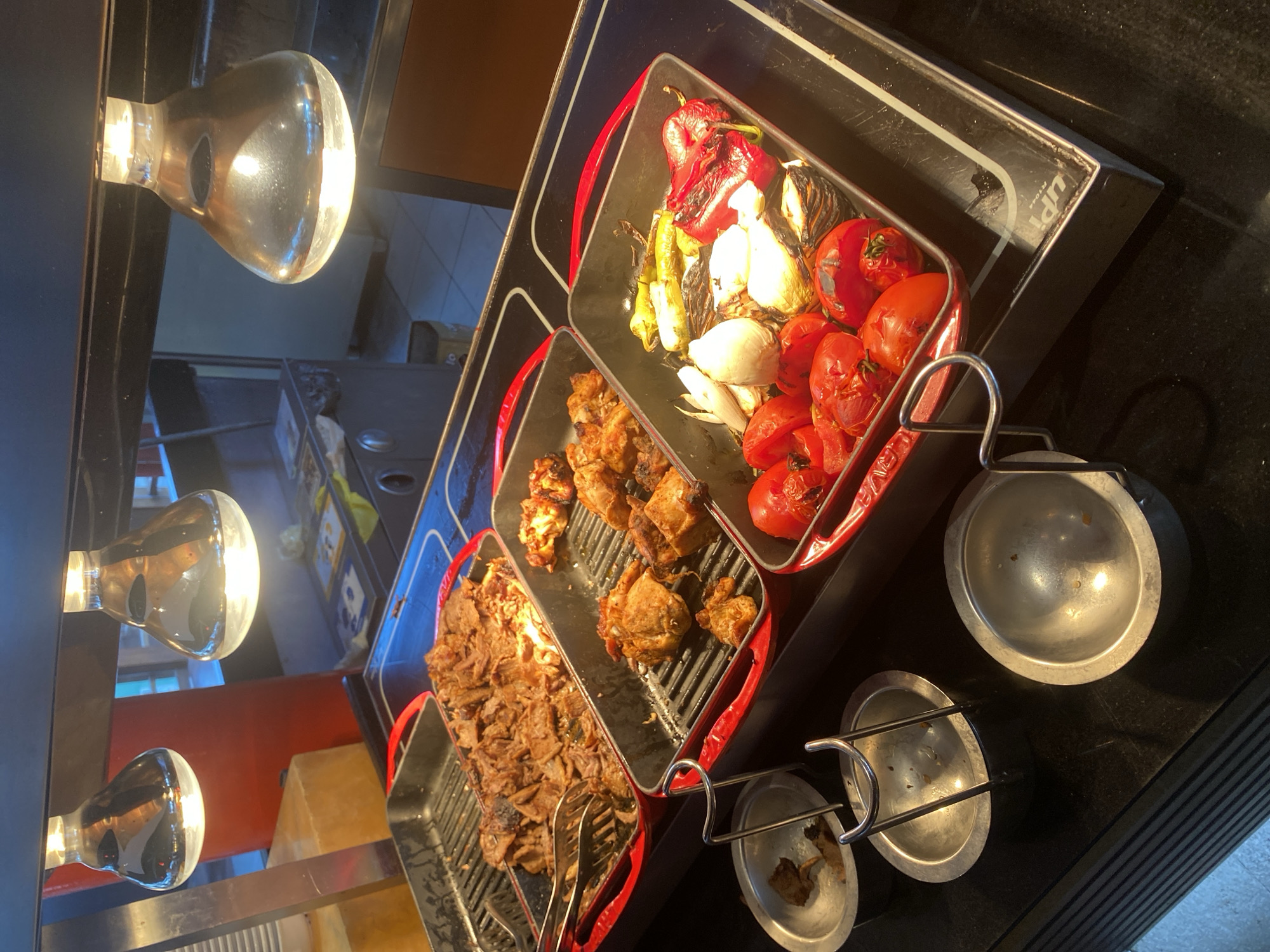 The image shows a buffet station with several dishes of cooked food. In the foreground, there are several small metal bowls, some of which appear to be empty or contain remnants of food. Behind the bowls, there are three rectangular metal trays containing food. The leftmost tray holds a pile of thinly sliced meat, likely some kind of roast or shawarma, with tongs placed in it. The middle tray contains pieces of what appear to be seasoned chicken. The rightmost tray holds a variety of grilled vegetables, including tomatoes, onions, and peppers. The entire setup is under heat lamps, indicated by the illuminated bulbs visible at the top of the frame. The buffet appears to be set up on a black countertop with metallic accents. The background shows a glimpse of a restaurant or food service area, with glimpses of walls and possibly other buffet elements. The lighting is warm and focused on the food. The scene suggests a food service environment where dishes are kept warm for self-service. Without further context or visible signage, it is not possible to definitively identify the specific location within Aksu, Turkey.