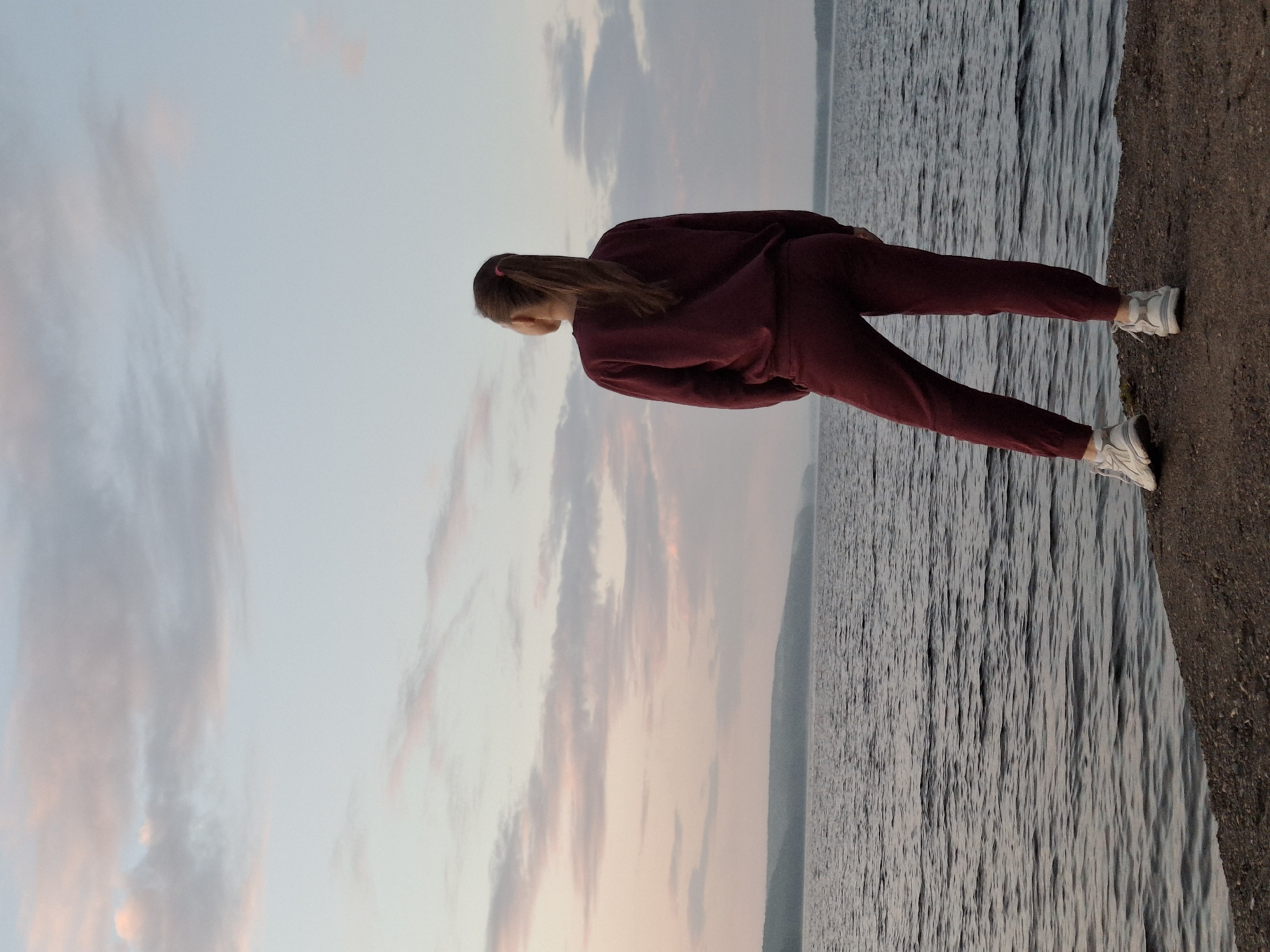 A person stands on the rocky shore of the Kama River, looking out at the water. The person is wearing a maroon tracksuit and white sneakers. The sky is a pale pink and blue, with scattered clouds. The water is calm and reflects the sky. In the distance, a hazy, tree-covered shore is visible. This location is near the city of Chaikovsky, Russia.