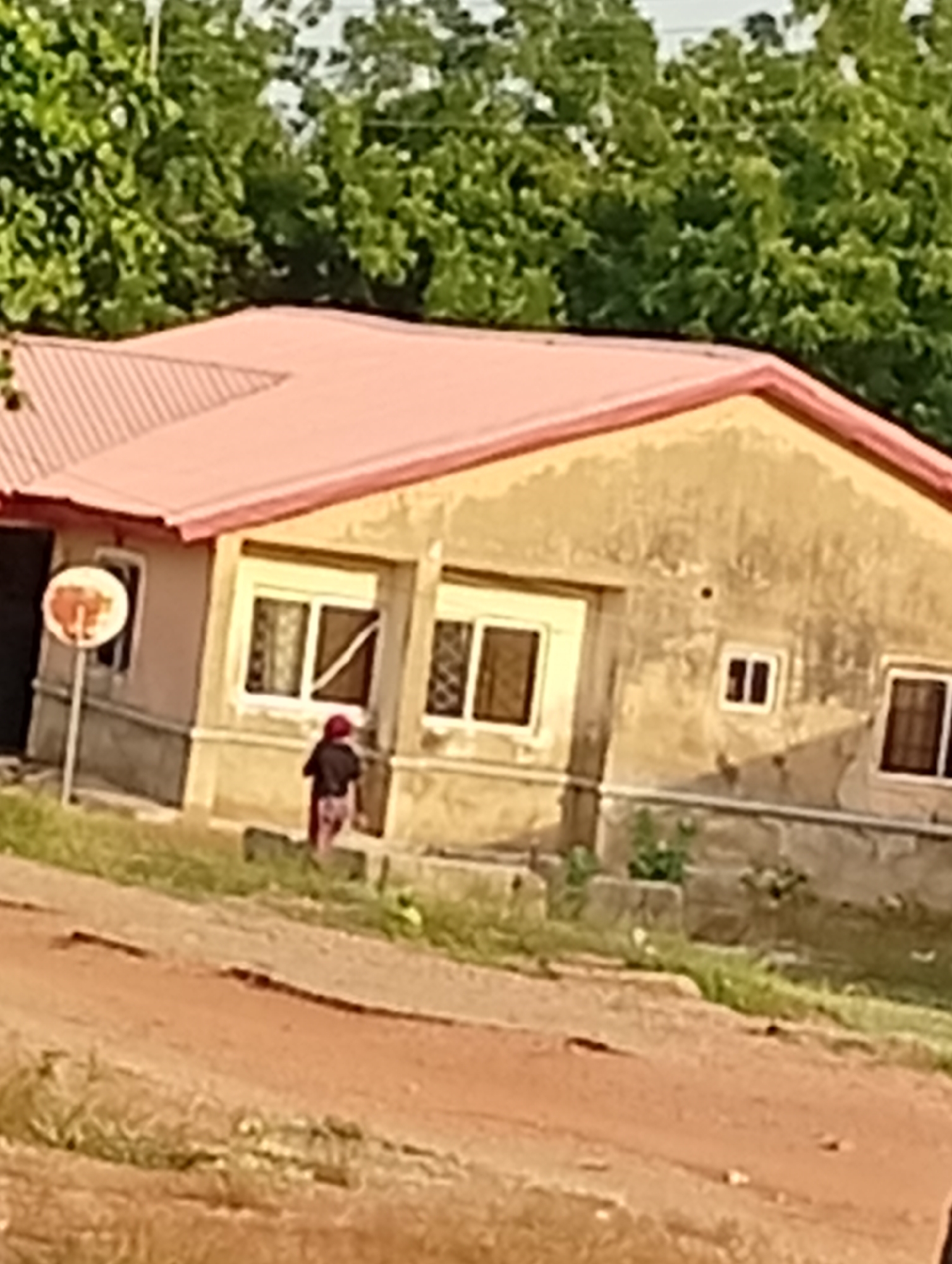 The image depicts a single-story building with a pinkish-red roof in an undefined area of Unknown, Unknown. The building is light brown with visible windows. A small, indiscernible figure of a person is visible in front of the building. In the foreground, there is a dirt road or area. Green foliage is visible in the background. A round sign is to the left of the building. The overall image is somewhat blurry and the angle is slightly off-center.