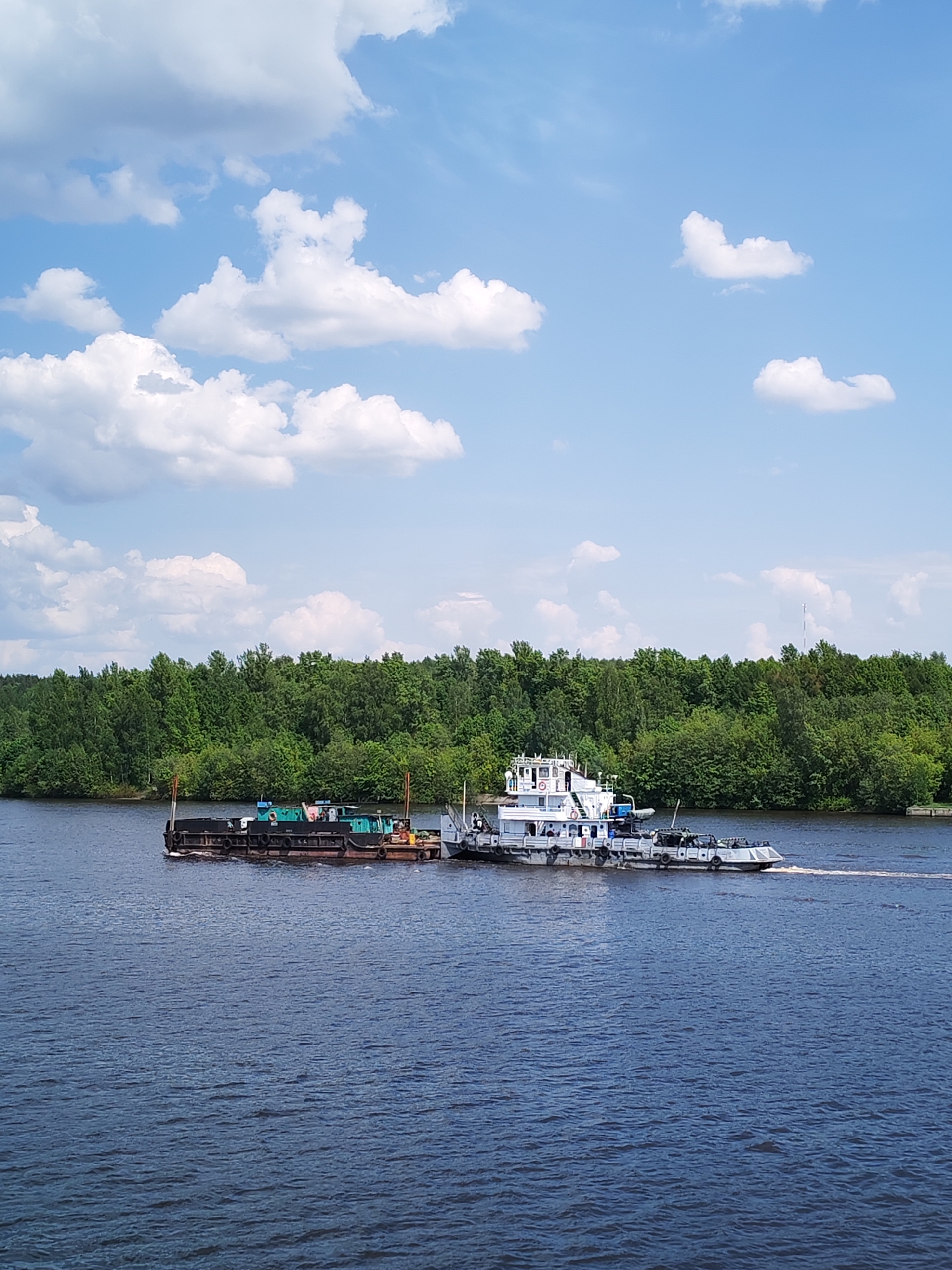 The image depicts a river scene in Ermolino, Russia. In the foreground, a dark blue river occupies most of the frame, its surface slightly rippled. A tugboat, predominantly white with blue accents, is connected to a darker, flat-decked barge, loaded with unspecified materials, and is moving across the river from left to right. The tugboat appears to be actively towing the barge. The river is flanked by a dense green forest on the opposite bank, providing a backdrop to the river traffic. Above, a bright blue sky is scattered with puffy white clouds.