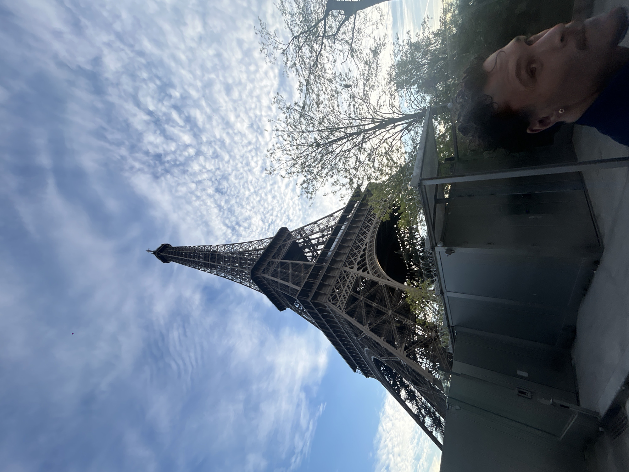 The image shows the Eiffel Tower in Paris, France, viewed from a low angle. The sky is blue with scattered white clouds. The top of the tower extends into the upper left portion of the image. A man is partially visible in the upper right corner of the image, with dark curly hair and wearing a blue shirt. A tree with bare branches is visible near the top of the frame. A gray structure is visible along the right side of the frame.