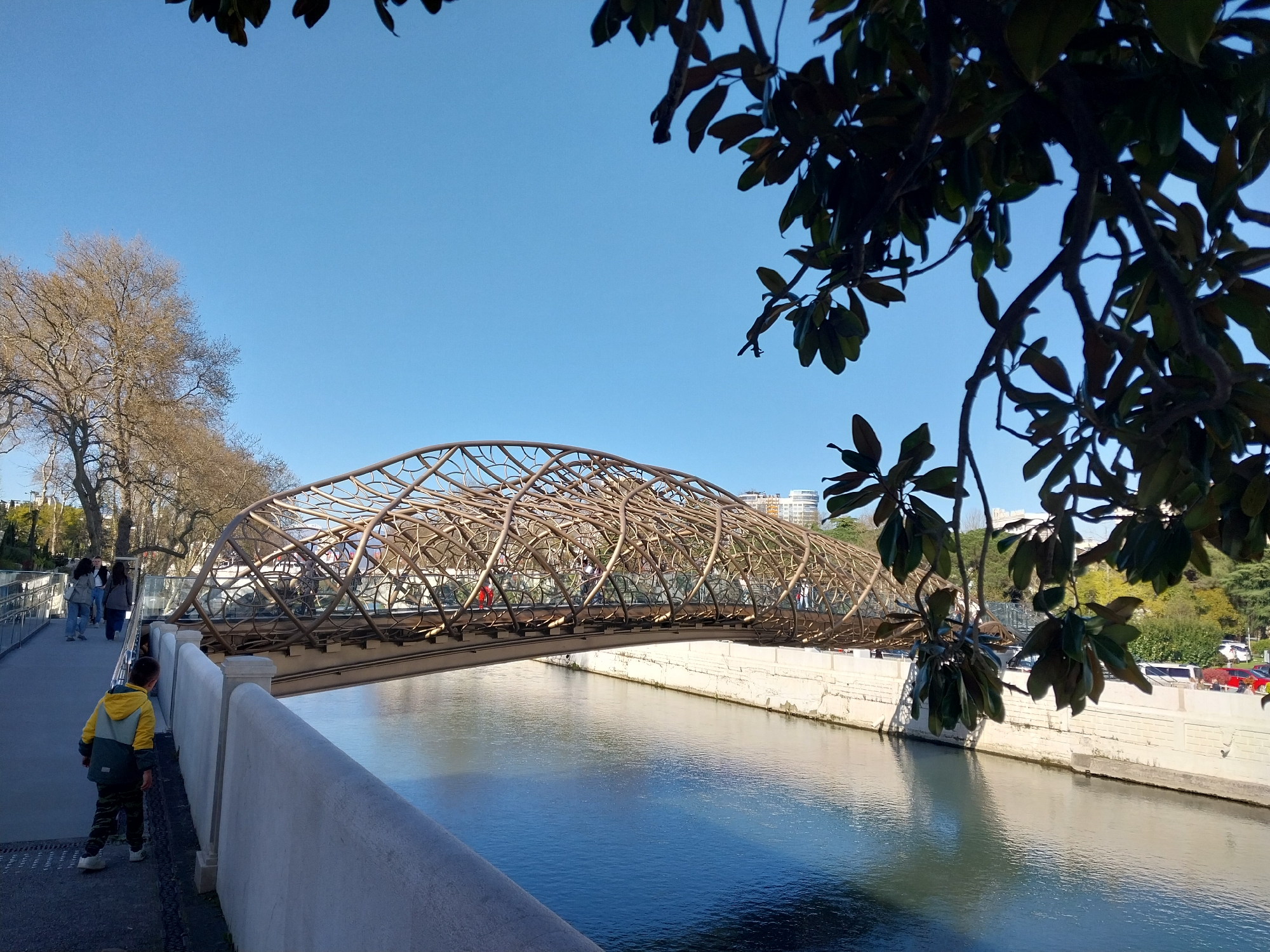 This image depicts a sunny outdoor scene in Sochi, Russia, featuring a distinctive pedestrian bridge spanning a tranquil river. In the foreground, a young child, dressed in a yellow and grey jacket with camouflage pants, walks along a paved pathway beside a low, white concrete wall.
The central focus of the image is the pedestrian bridge, which features a unique, intricate lattice structure made of what appears to be metallic, possibly bronze or gold-colored, material. This decorative, arching framework extends over the river, and several people can be seen walking across it. The river below reflects the clear blue sky and offers glimpses of the riverbed or deeper water.
On the left side of the river, bare trees line the bank, their branches silhouetted against the bright sky, suggesting either late autumn or early spring. The right side of the image features lush green foliage and distant buildings, indicating an urban setting. Overhead, in the upper right and across the top, dark green leaves from an overhanging tree branch partially frame the scene.
The clear, vibrant blue sky and strong natural lighting indicate a bright, sunny day. The scene conveys a sense of calm and leisure, with individuals enjoying a stroll by the river and across the architecturally interesting bridge. There is no visible text in the image.
