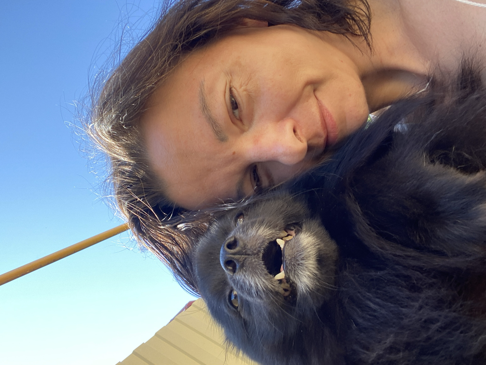 A close-up selfie captures a woman and a black Pomeranian dog against a clear blue sky and a beige building. The woman, on the right, has dark brown hair and is smiling slightly. The dog, on the left, is looking directly at the camera with its mouth open, revealing its teeth and pink tongue. The image appears to have been taken outdoors in a sunny location, possibly Yuzhny, Russia, given the context.