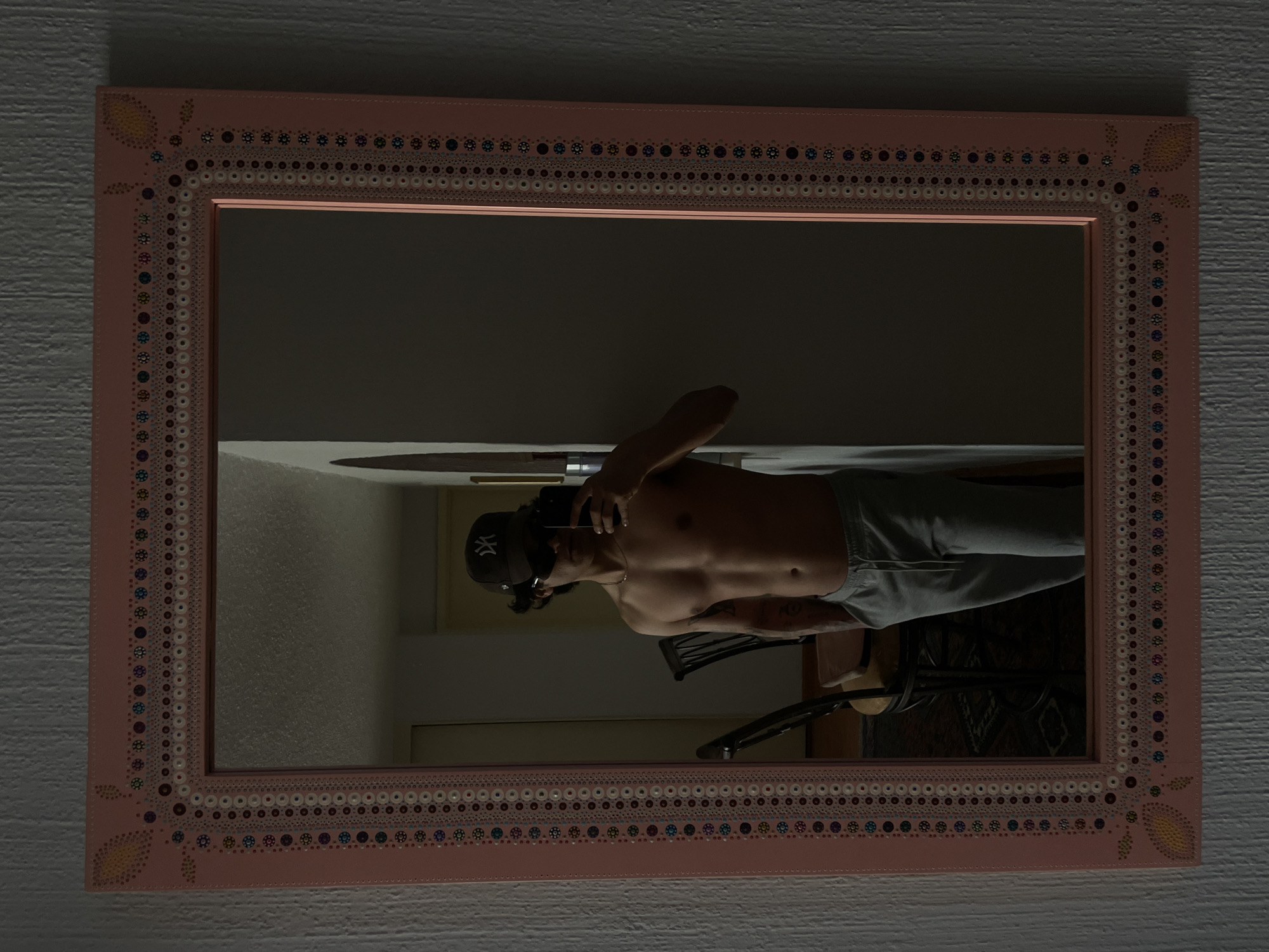 The media file displays a mirror selfie taken indoors. A large, rectangular mirror with a wide, light pink frame is centered in the image. The frame is ornately decorated with intricate patterns of small, colorful dots in various shades of blue, purple, white, and brown, arranged in concentric circles and lines. Stylized yellow leaf or flower motifs adorn each corner of the frame. The mirror is mounted on a textured, light gray wall.
Reflected in the mirror is a shirtless person, visible from the waist up, showcasing an athletic build. They are wearing light gray sweatpants or shorts and a dark baseball cap with a prominent white "NY" logo on the front. Their right hand is raised near their head, holding what appears to be a phone, suggesting they are capturing the mirror selfie. The person's face is largely obscured by the cap and the phone.
The background reflected behind the person is a dimly lit interior room with light-colored walls. Portions of a dark chair or piece of furniture and a patterned rug or mat are visible below the person's reflection. The overall lighting is low, indicative of an indoor setting with either artificial light or limited natural light. Based on the provided context, the scene is likely a home in Mexico City, Mexico. No specific emotions are discernible due to the obscured face.
