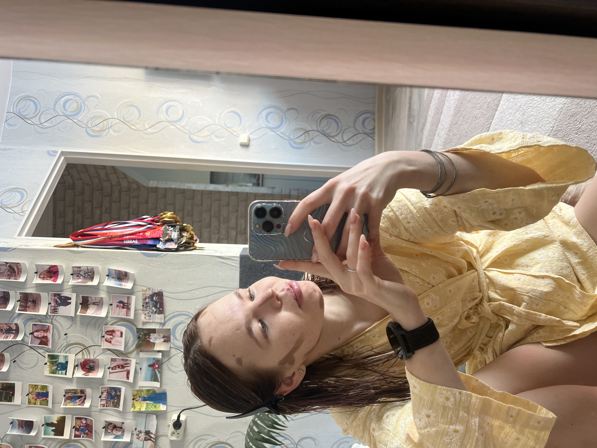 A young woman is depicted taking a mirror selfie in a residential interior in Chaikovsky, Russia. She has dark, wet hair, pulled back on the left side with a black clip, and makeup contouring is evident with darker streaks on her cheeks and forehead. She is wearing a yellow robe adorned with a white daisy pattern. On her left wrist, she wears a black smartwatch, and on her right hand, a ring is visible on the ring finger, along with three thin bracelets on her right wrist. She holds a smartphone, appearing to be an iPhone, with both hands to capture her reflection. The background features a wall decorated with numerous small, printed photographs arranged in a dense grid, an electrical outlet, and wallpaper displaying a light base with swirling lines and blue and gold circular motifs. To the right, a doorway framed in light wood leads to an adjacent room with light-colored brick-patterned wallpaper. A cluster of sports medals with red, white, and blue ribbons hangs on a hook near the doorway. A light-colored carpet or rug covers the floor, visible in the mirror's reflection.