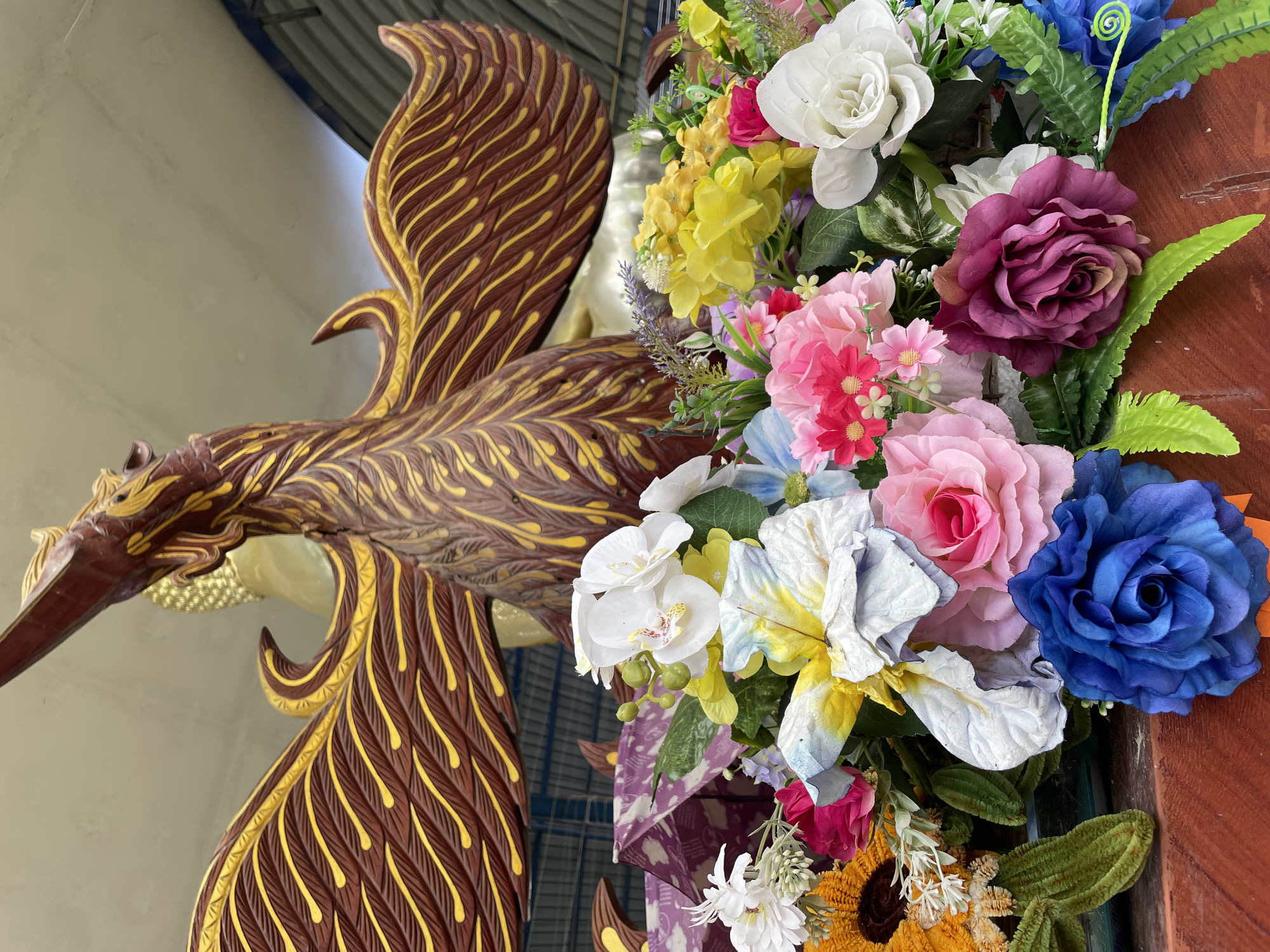 A close-up, eye-level shot focuses on a vibrant artificial flower arrangement in Tambon Na Chom Thian, Thailand. The bouquet, positioned on a reddish-brown wooden surface, bursts with a variety of colorful blooms, including pink roses, white orchids, yellow flowers, and a striking blue rose. Green foliage and some decorative elements like a purple patterned material are also visible within the arrangement. Behind the flowers, a large, ornately carved wooden statue of a mythical bird, possibly a phoenix, with spread wings dominates the upper portion of the image. The bird is predominantly brown with gold accents on its feathers and wing patterns. In the background, partially obscured by the bird statue, is a glimpse of a golden, serene Buddha statue, suggesting a religious or cultural setting. The environment appears to be indoors or under a covered structure, with a light-colored ceiling and hints of metal roofing visible in the upper right corner. The lighting is bright, casting subtle shadows. No people are present in the scene.