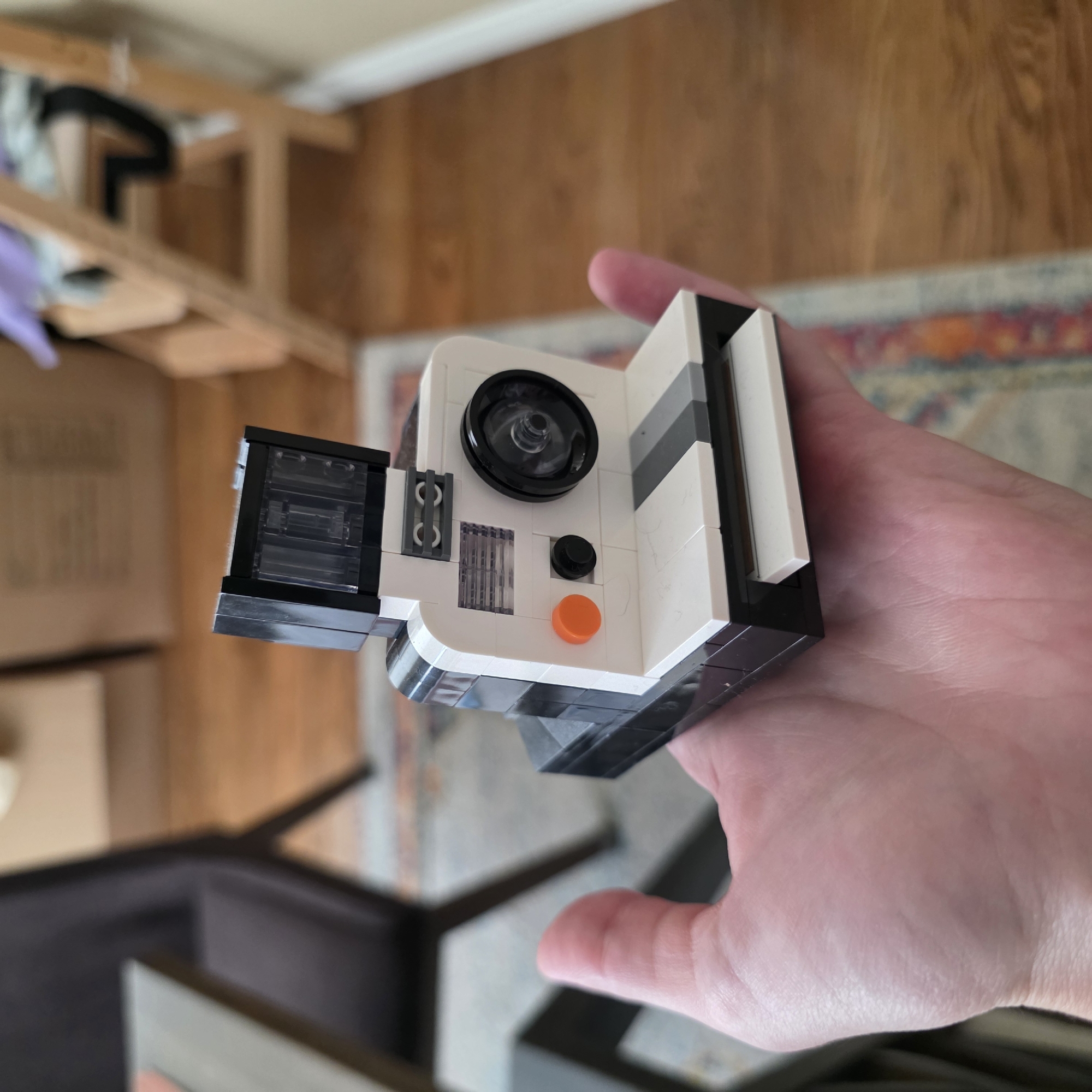 An extreme close-up shows a person's hand holding a LEGO model of a Polaroid camera. The camera is predominantly white with black accents, featuring a clear plastic flash unit at the top, a lens, and various buttons and details. The hand is pale pink and appears to be supporting the camera from underneath. In the background, out of focus, are wooden floorboards, a portion of a rug with geometric patterns in muted colors, and the legs of a wooden chair. The setting suggests an indoor environment, possibly a home. There is no indication of the city of Portland, United States of America.